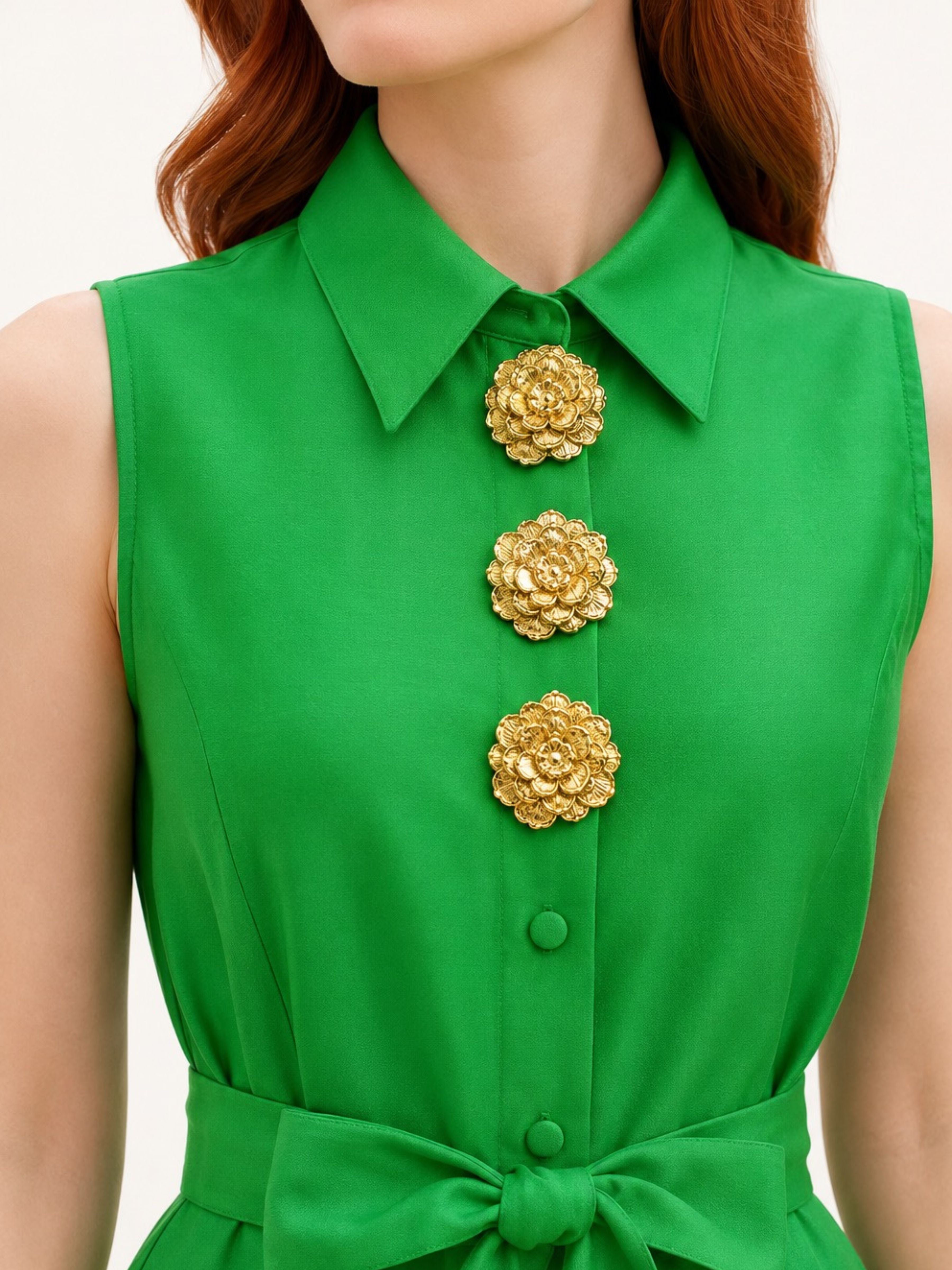 A close-up of a woman wearing a bright green sleeveless collared dress or top with three large textured gold flower-shaped buttons down the front and a matching waist tie.