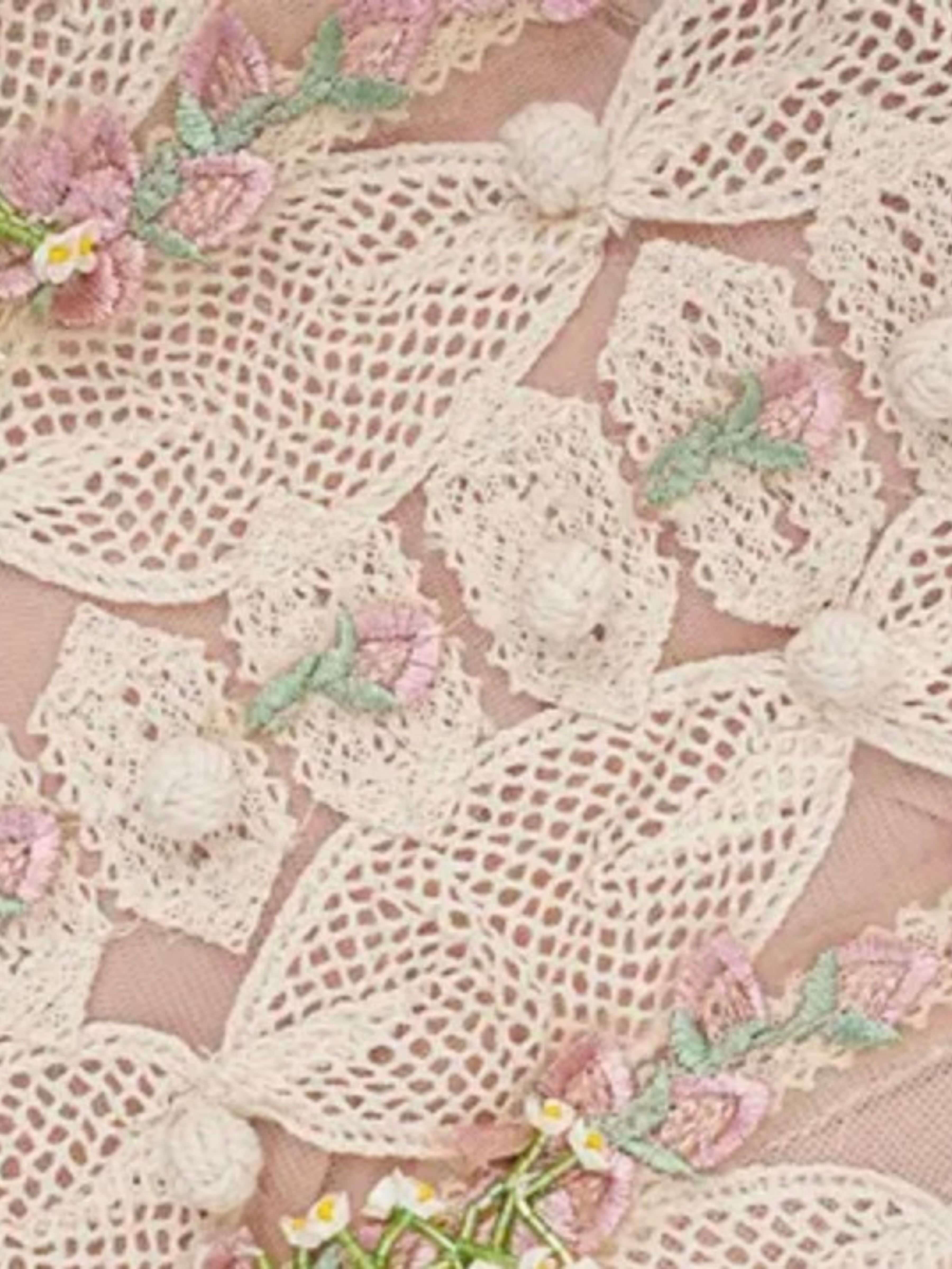 Close-up of a delicate cream lace fabric with embroidered pink flowers, green leaves, and small white pom-pom accents on a soft blush pink background.