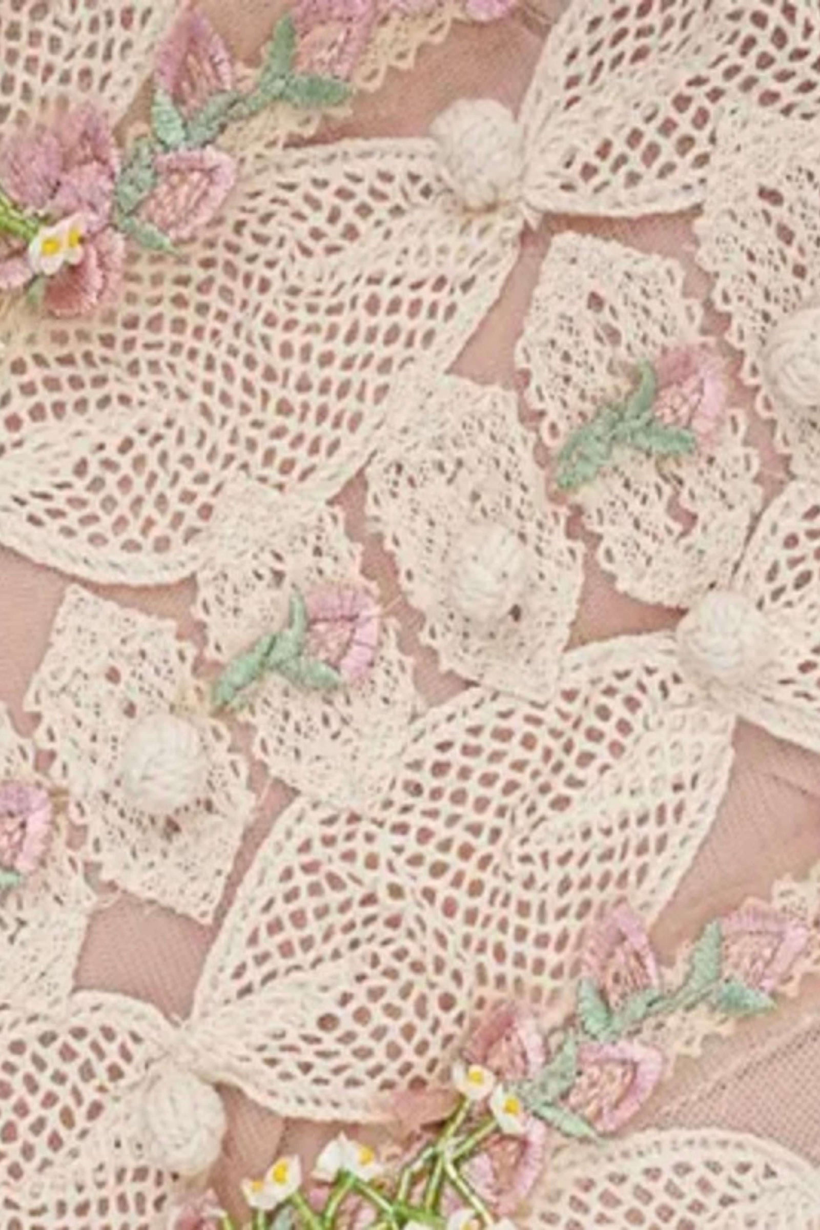 Close-up of a delicate cream lace fabric with embroidered pink flowers, green leaves, and small white pom-pom accents on a soft blush pink background.
