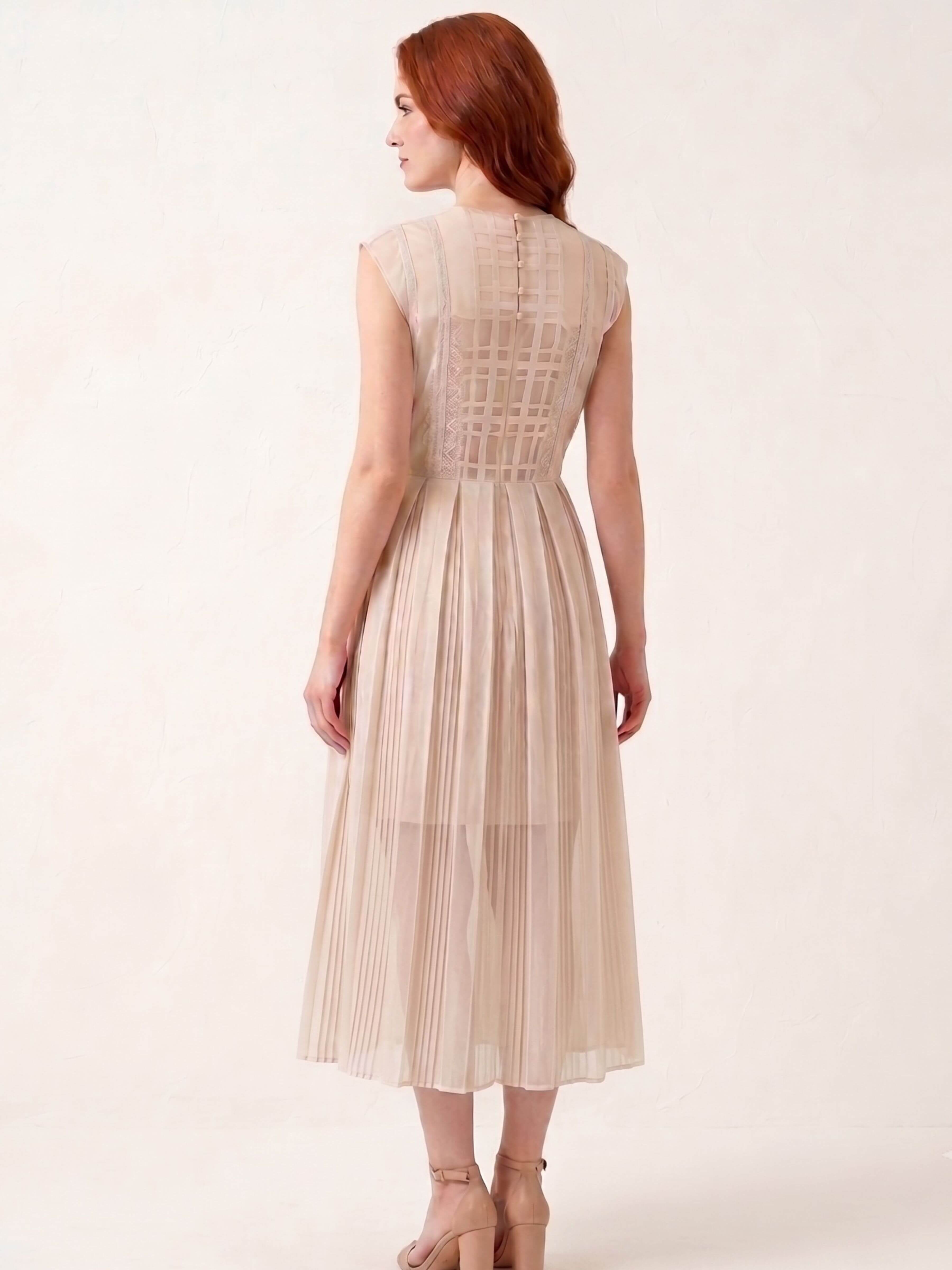 A woman with red hair is shown from the back wearing a sleeveless, pleated midi dress in a light beige or champagne color. The dress features a structured, crisscross lace-like pattern across the upper back and a pleated skirt.