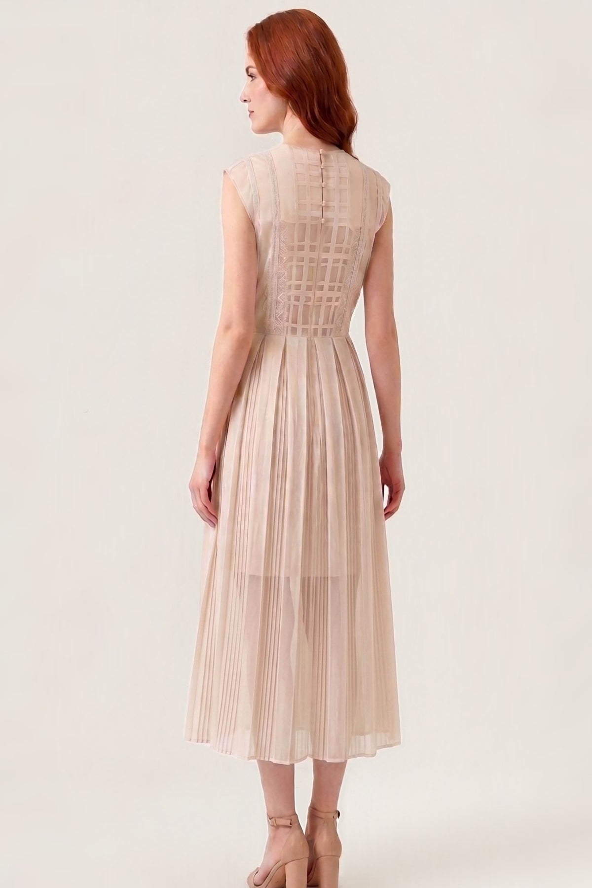 A woman with red hair is shown from the back wearing a sleeveless, pleated midi dress in a light beige or champagne color. The dress features a structured, crisscross lace-like pattern across the upper back and a pleated skirt.