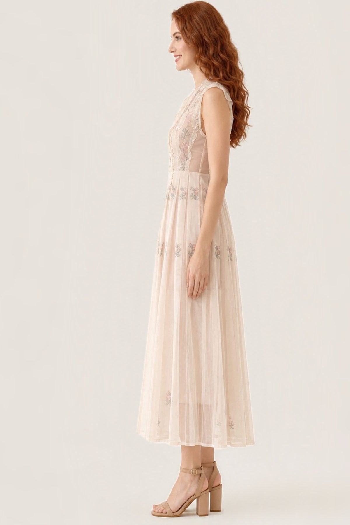 A woman in a sleeveless, floor-length cream tulle dress with floral embroidery and sheer lace bodice, paired with beige block-heel sandals, shown in profile against a warm off-white studio background.
