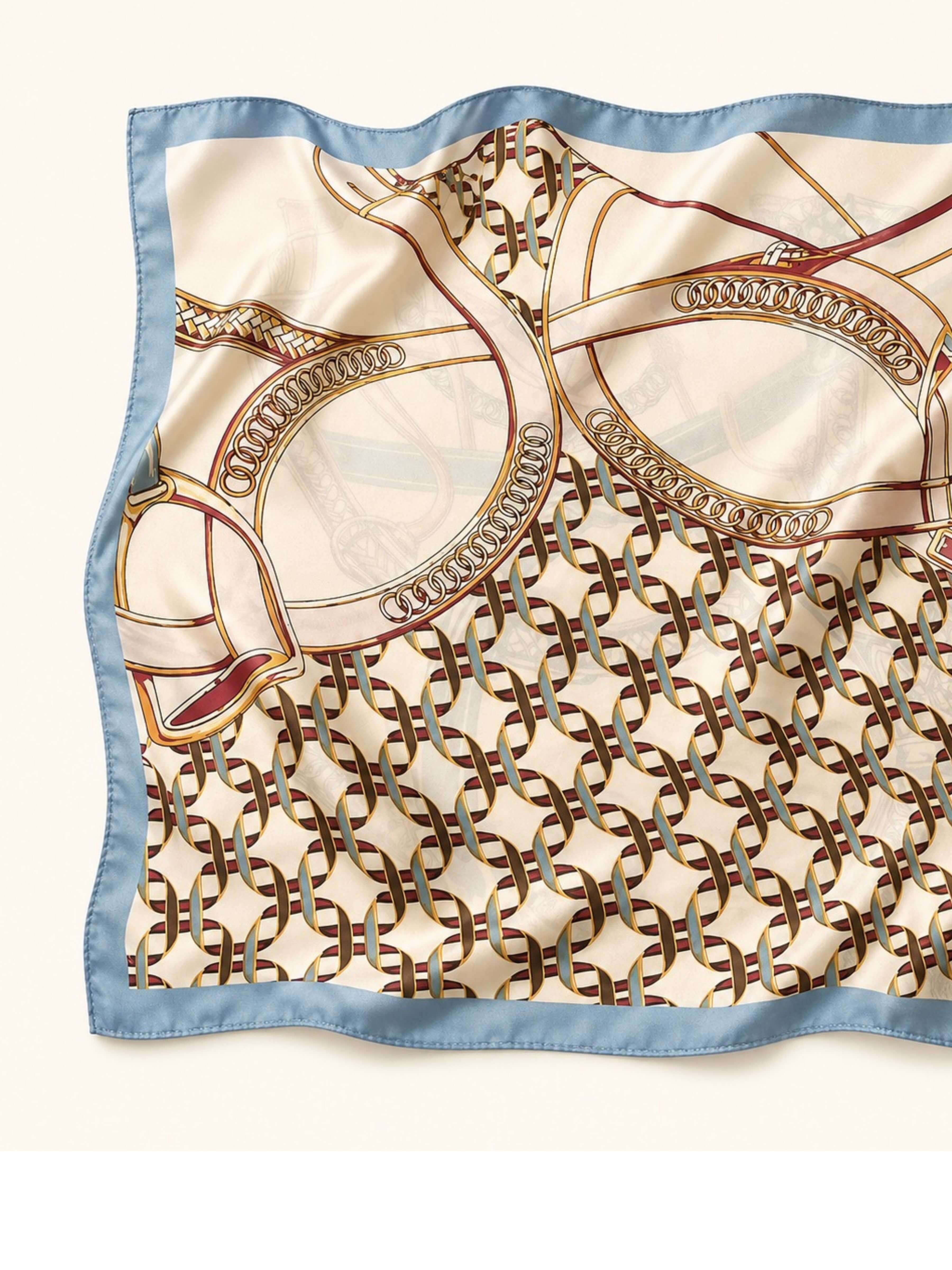 A square silk scarf with a light blue border, featuring an elegant cream and gold horse-and-carriage style pattern with brown interlocking chain motifs and subtle red accents, laid flat on a light beige background.