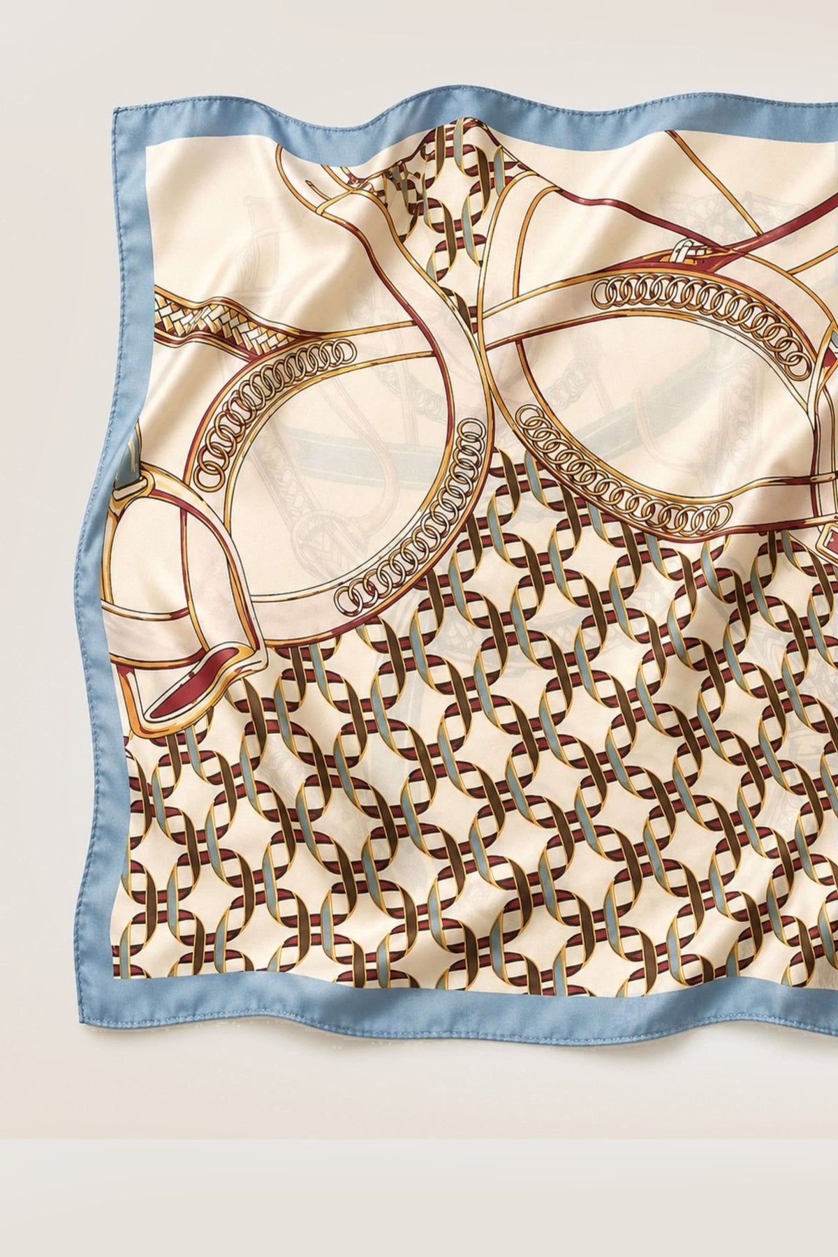 A square silk scarf with a light blue border, featuring an elegant cream and gold horse-and-carriage style pattern with brown interlocking chain motifs and subtle red accents, laid flat on a light beige background.