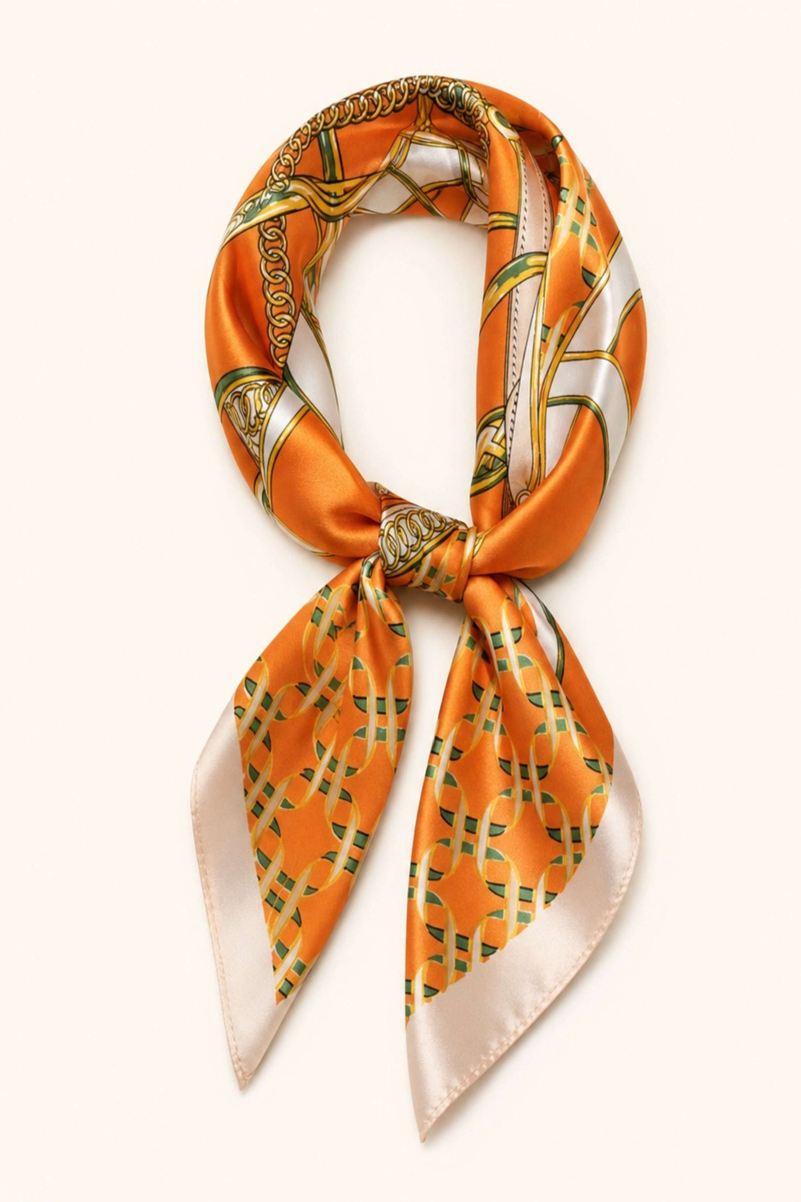 An orange silk scarf with a green and gold geometric pattern and cream borders, tied in a knot and displayed on a light cream background.