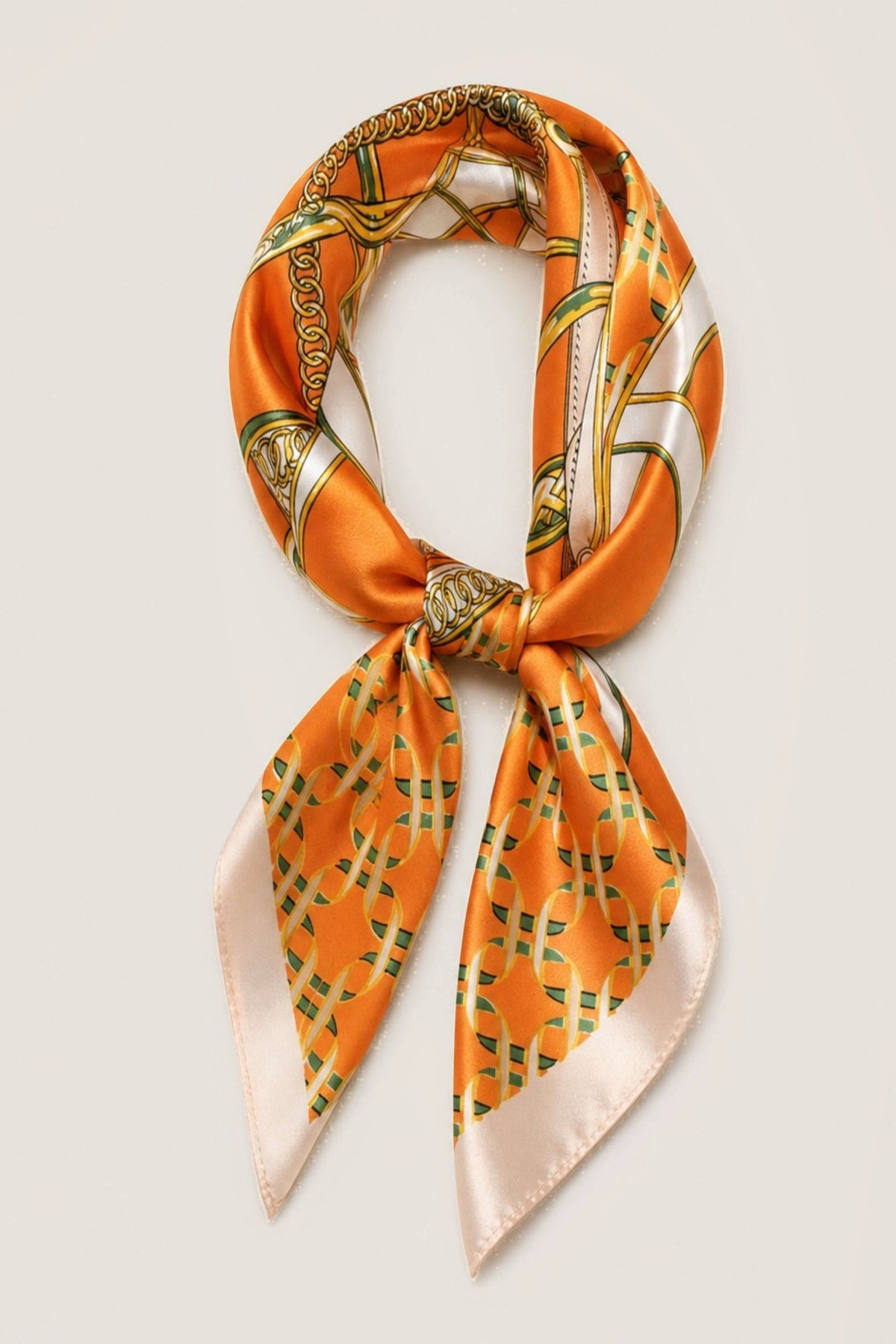 An orange silk scarf with a green and gold geometric pattern and cream borders, tied in a knot and displayed on a light cream background.