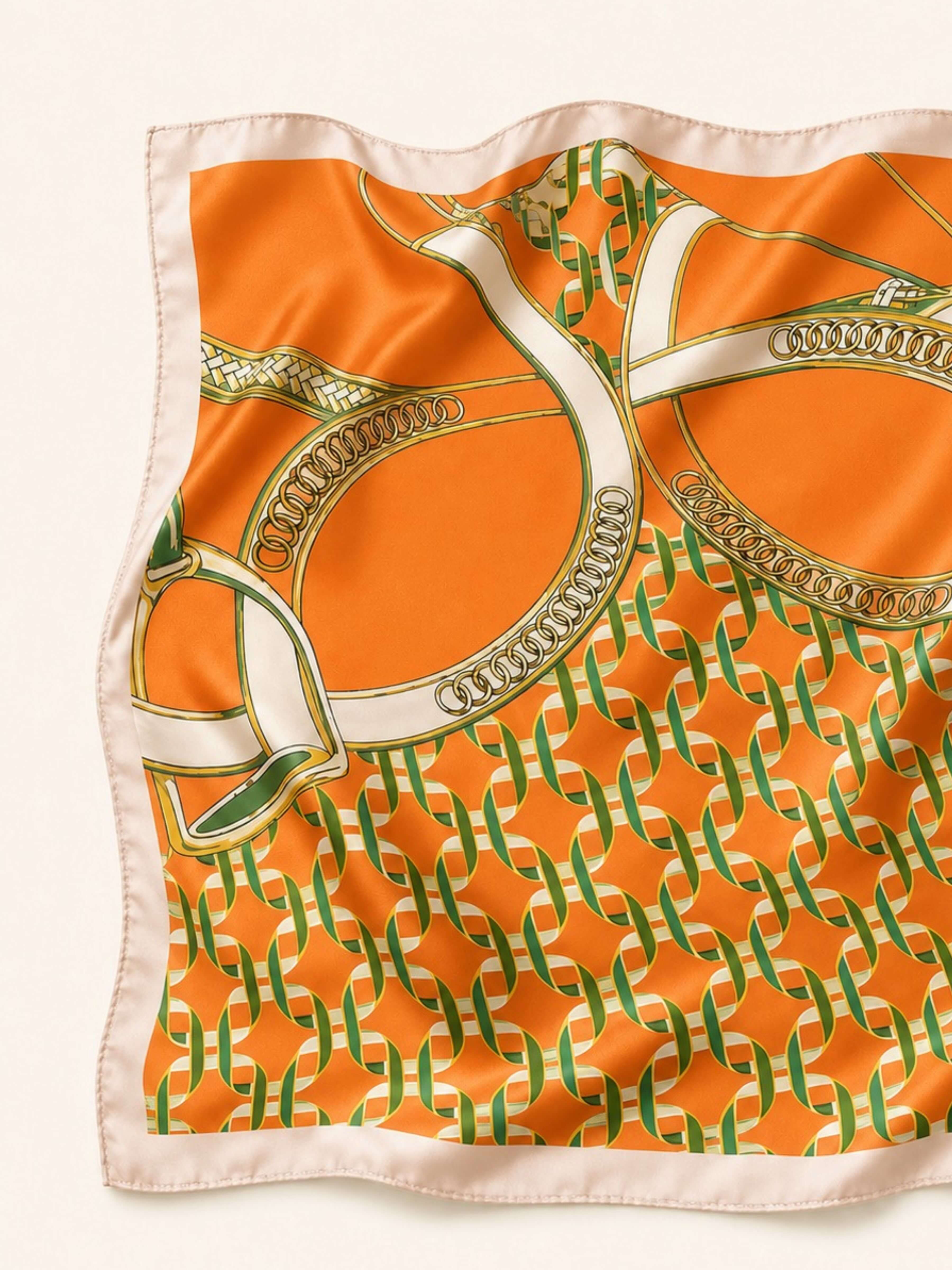 An orange silk scarf featuring a green and gold interlocking monogram pattern and a cream border, laid flat on a light beige background.