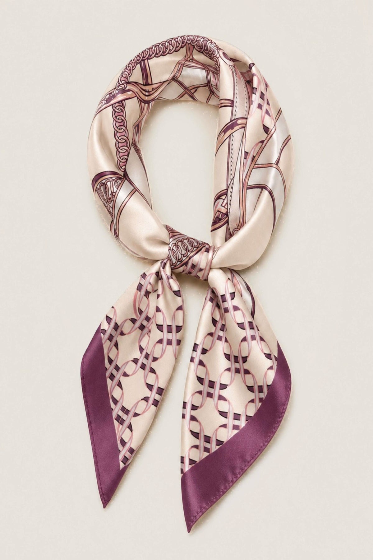 A silk scarf with a geometric interlocking chain pattern in purple and beige, tied in a knot and displayed against a light cream background.