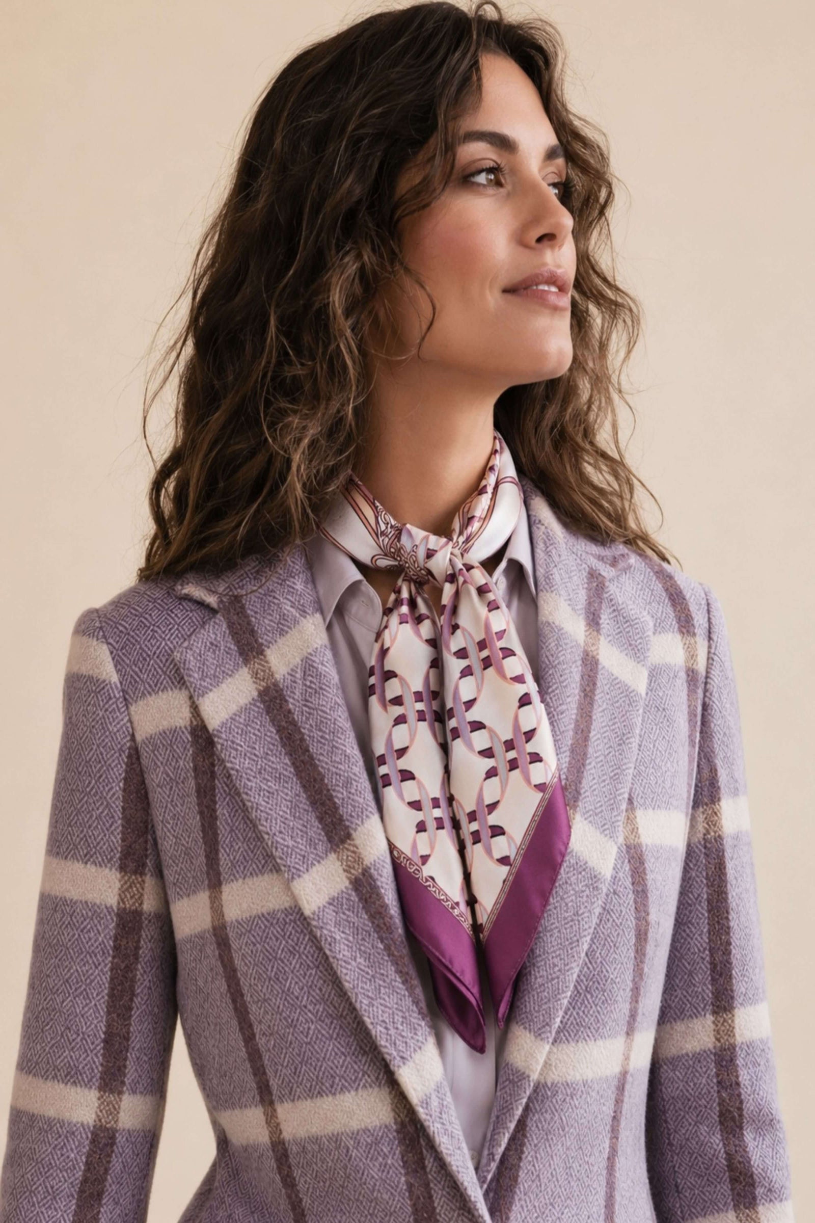 A woman wearing a lavender and beige plaid blazer with a patterned purple and white neck scarf, set against a solid beige background.
