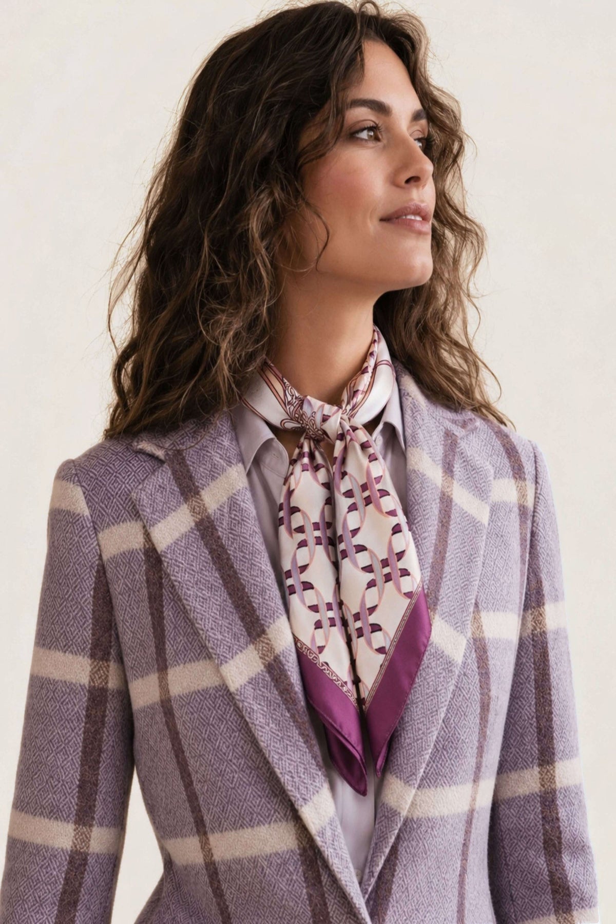 A woman wearing a lavender and beige plaid blazer with a patterned purple and white neck scarf, set against a solid beige background.