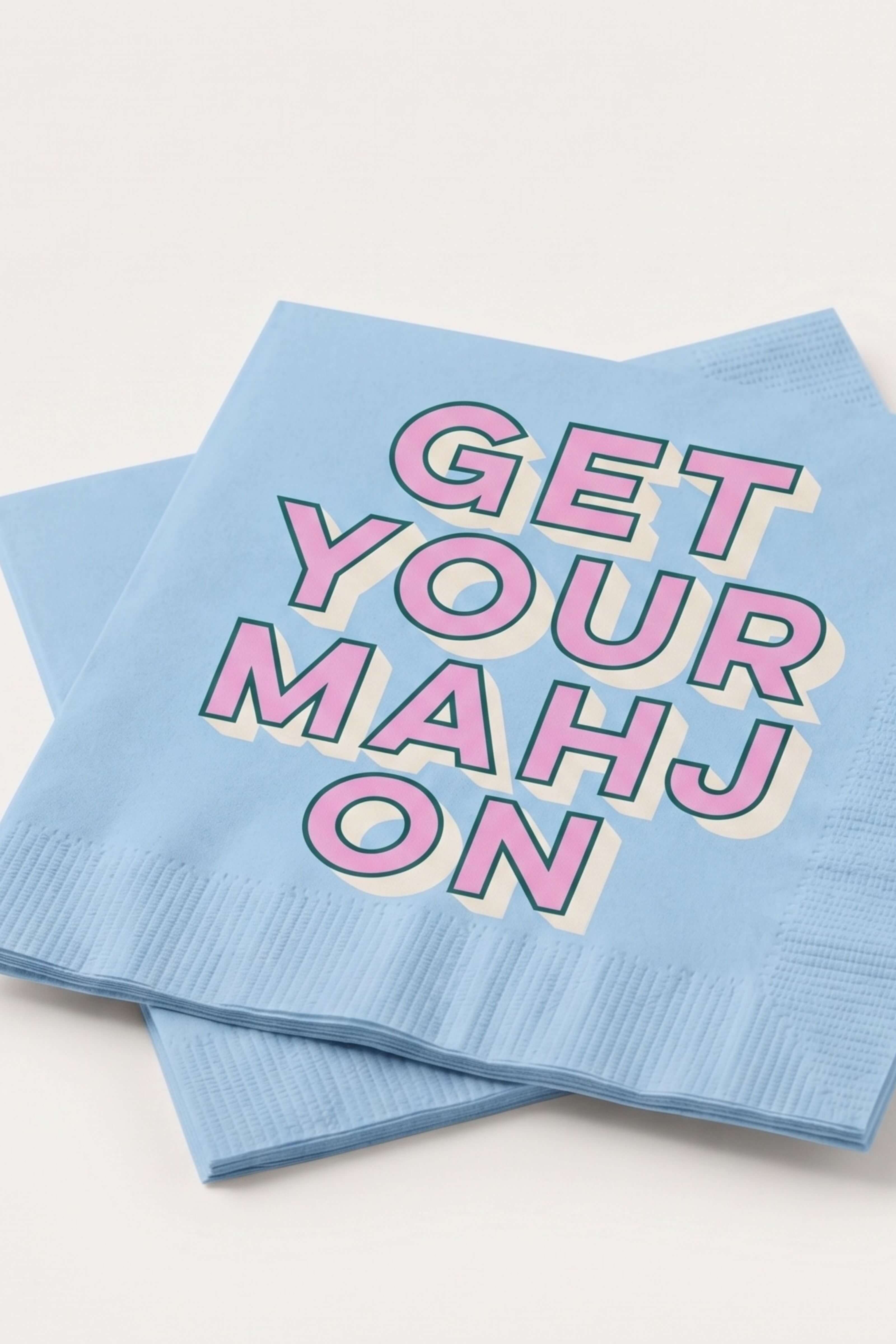 Two light blue paper napkins stacked on a white background, printed with the phrase 'GET YOUR MAHJON' in pink letters with a cream and dark outline.