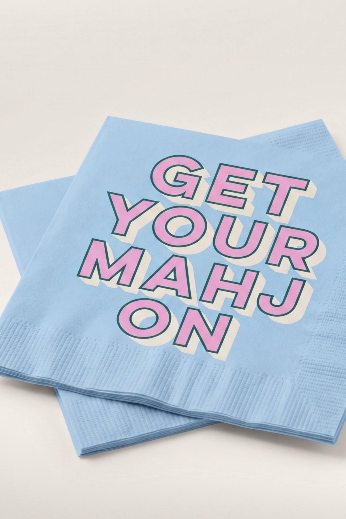 Two light blue paper napkins stacked on a white background, printed with the phrase 'GET YOUR MAHJON' in pink letters with a cream and dark outline.
