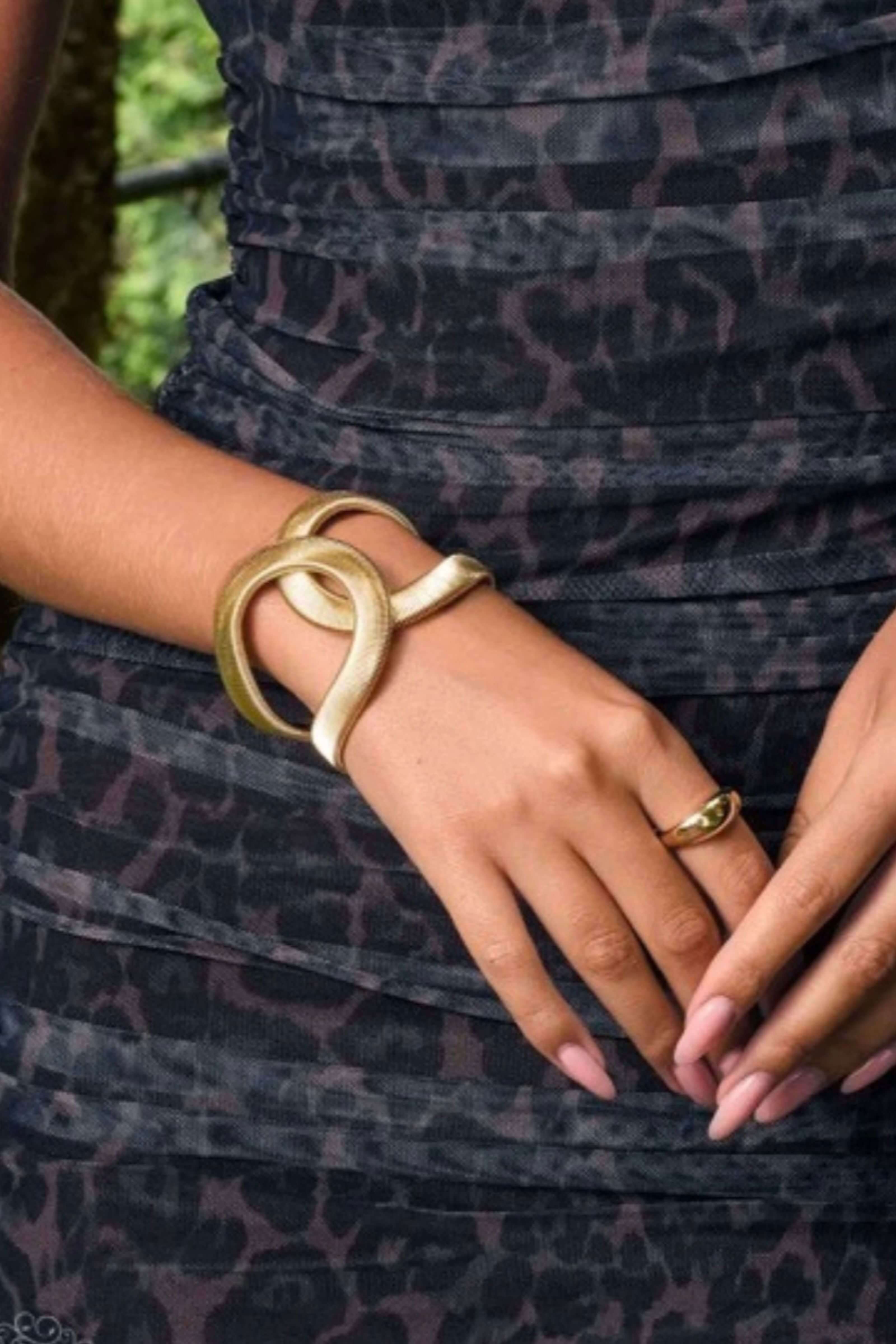 A close-up of a person's hands and torso wearing a chunky, double-looped gold bangle and a matching gold ring, set against a dark leopard-print dress.