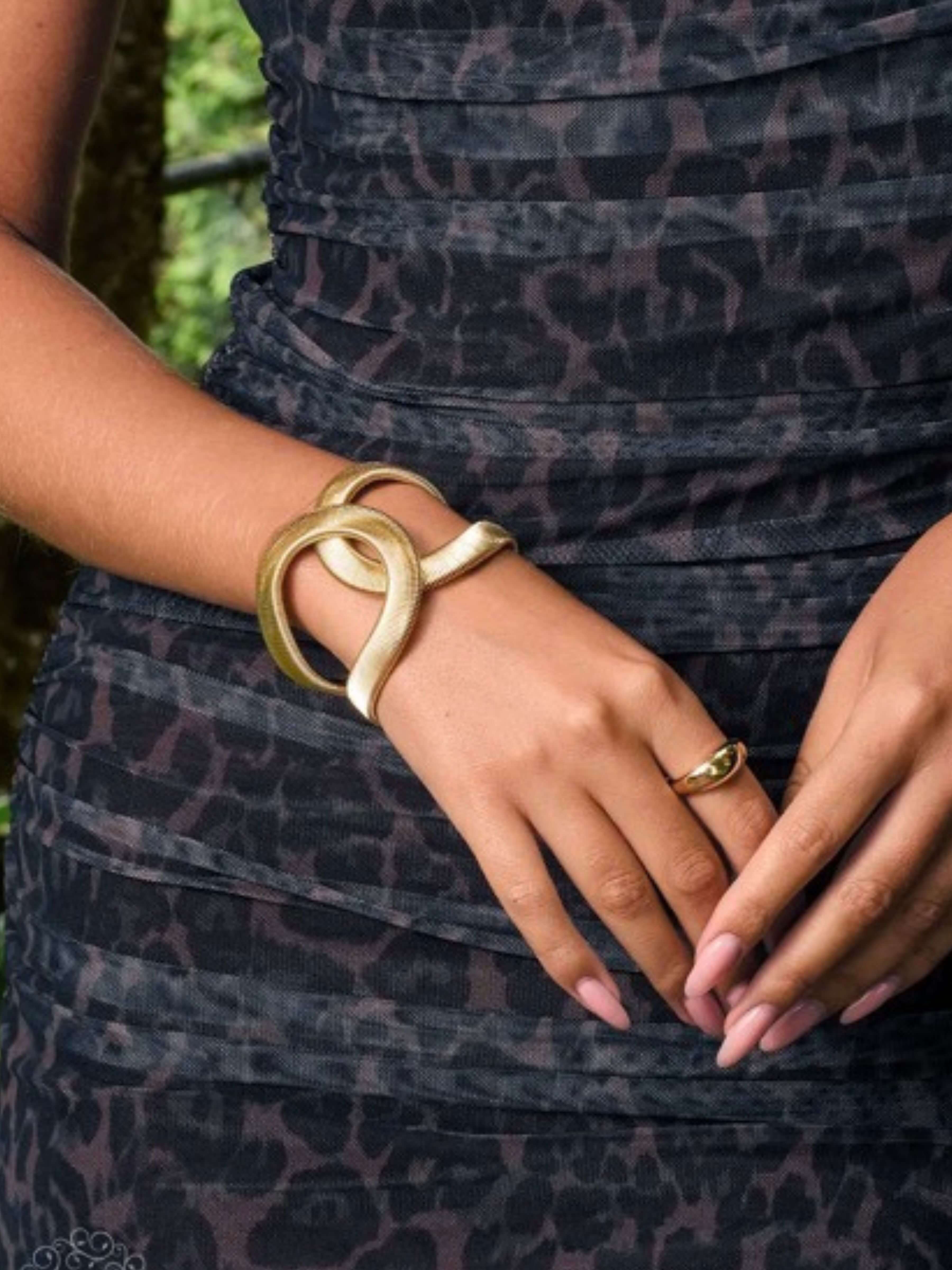A close-up of a person wearing a chunky gold wrap bracelet on one wrist and a matching gold ring on the other hand, styled with a dark leopard-print dress.