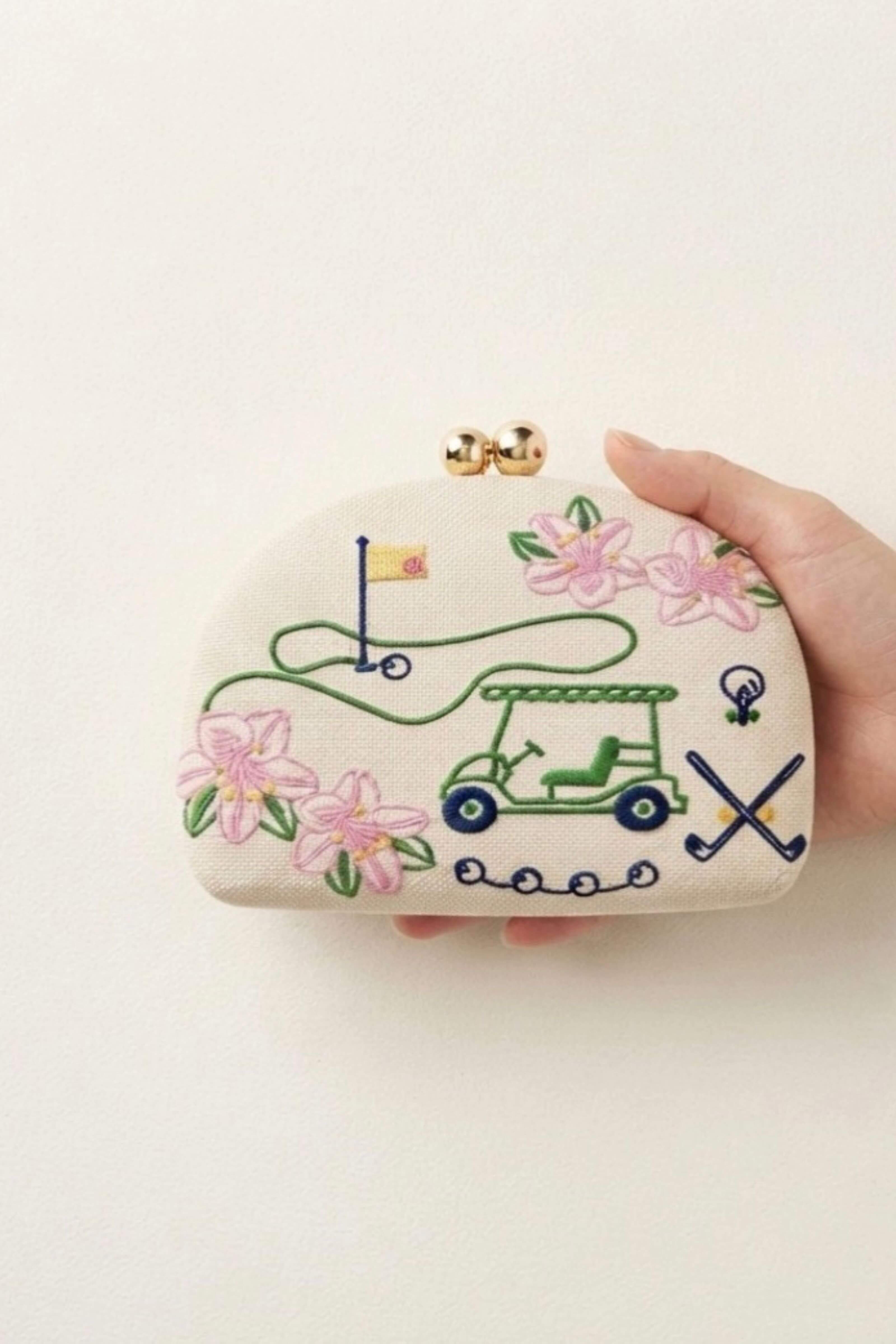 A hand holding a small cream-colored clutch purse decorated with embroidered golf-themed motifs, including a green golf cart, pink flowers, a flagstick, and crossed clubs, with a gold-tone clasp closure.