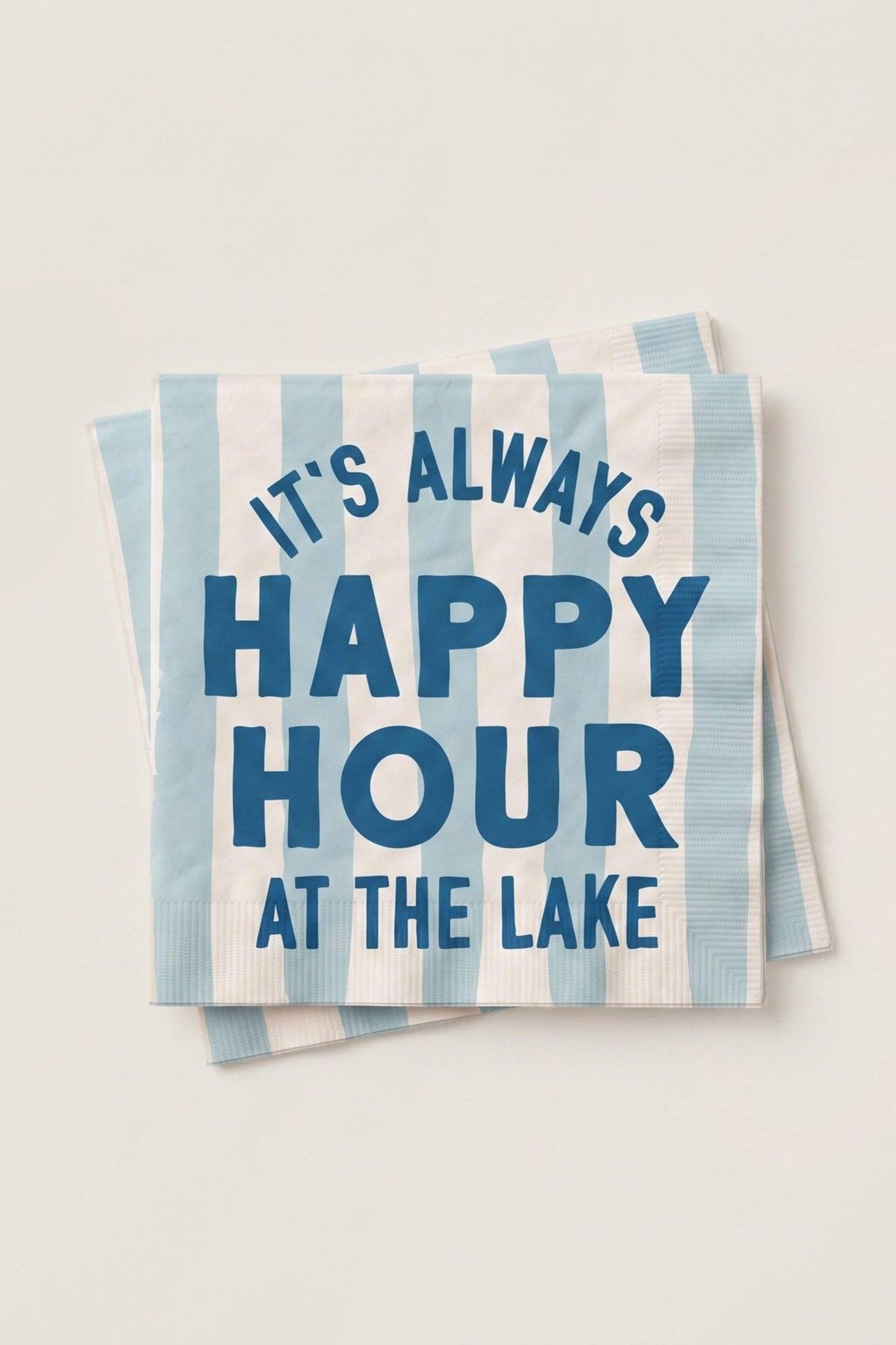 A stack of square paper napkins with light blue and white vertical stripes, featuring the text 'IT'S ALWAYS HAPPY HOUR AT THE LAKE' in dark blue lettering, set against a plain off-white background.