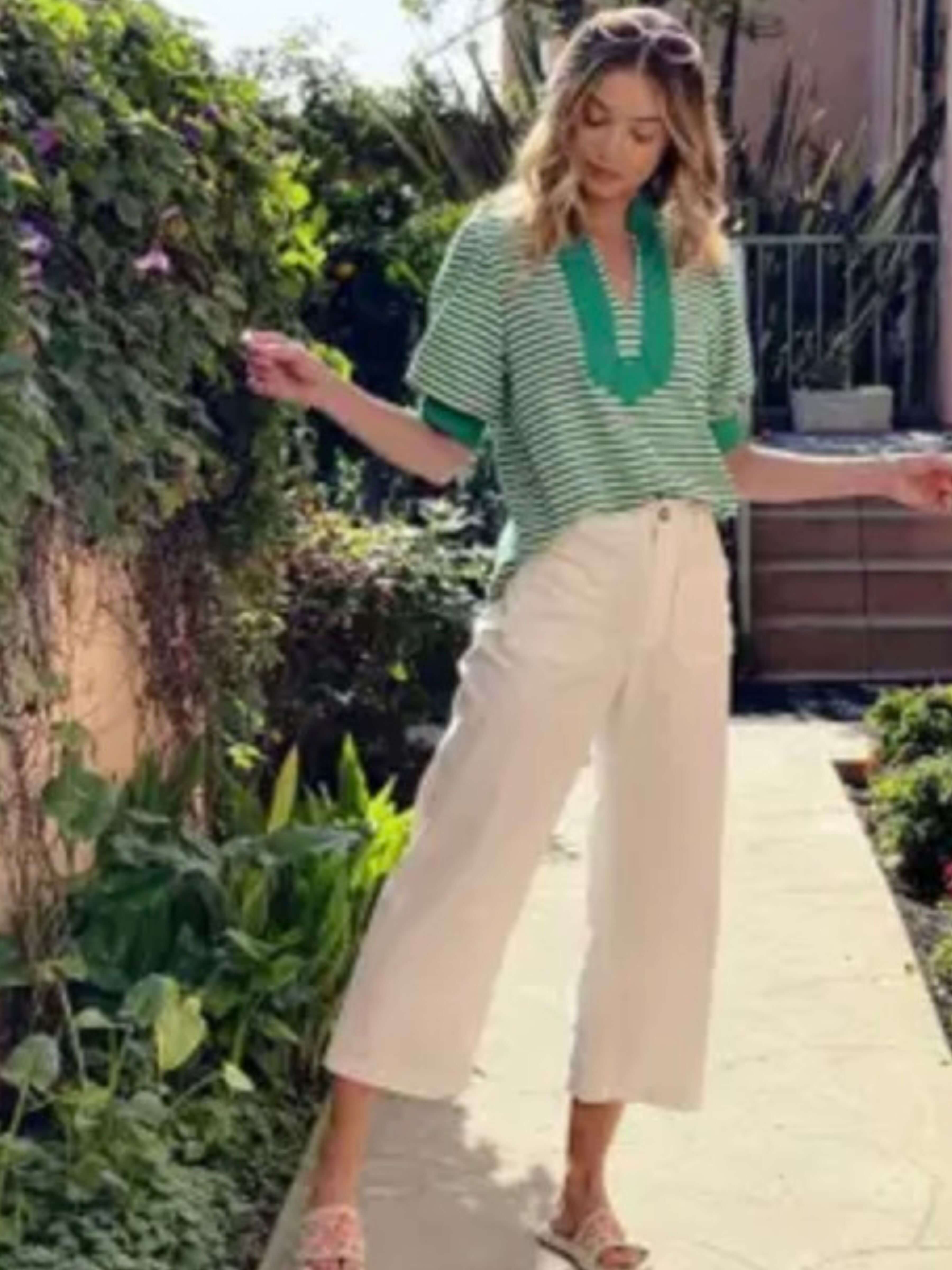 A woman posing outdoors on a stone pathway wearing a green and white striped short-sleeve top with a green V-neckline, paired with cream-colored cropped pants.