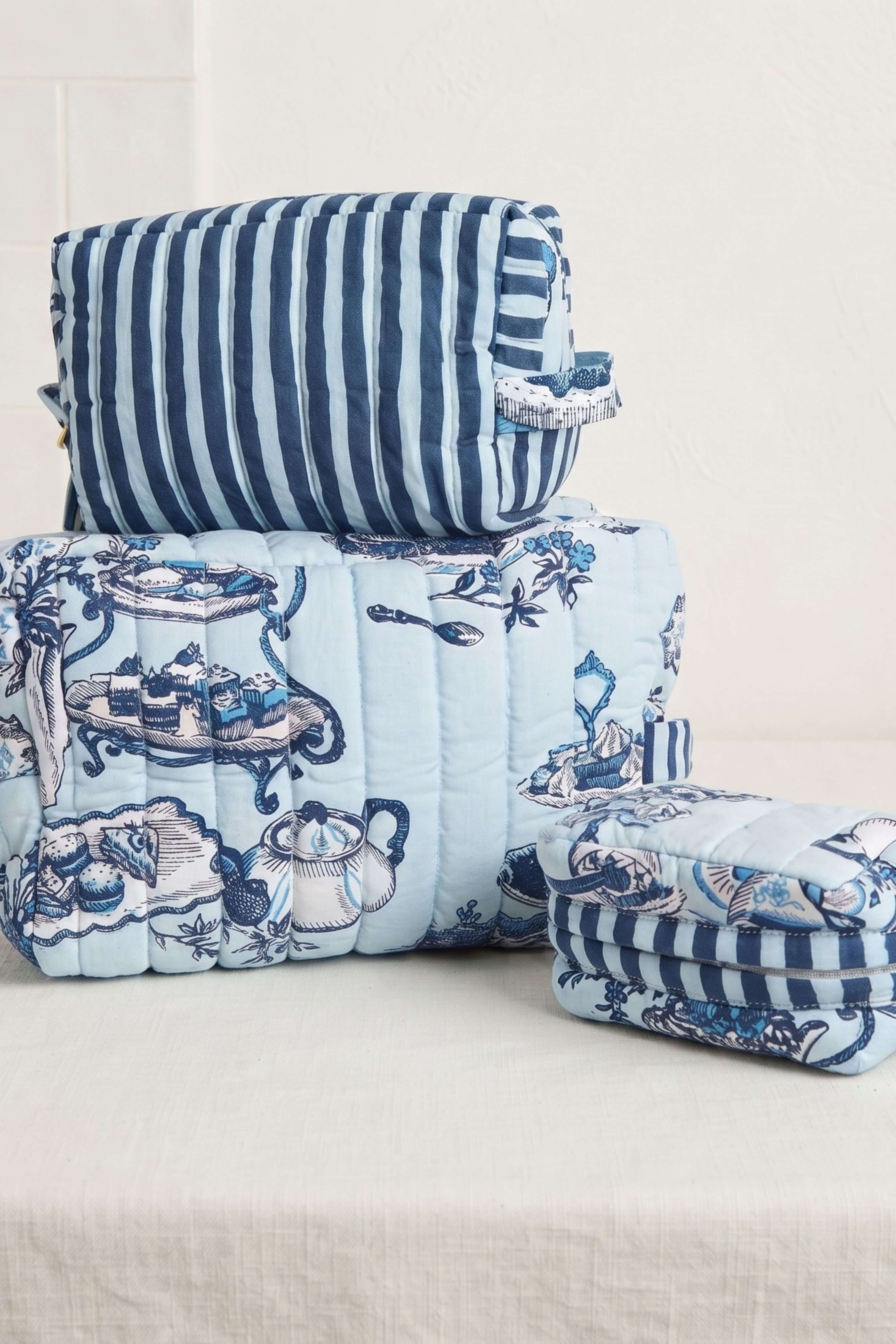 A coordinated bedding or textile set displayed on a light neutral surface, featuring a light blue quilted cover with a blue toile-style print, a striped pillow or cushion in navy and white, and folded matching striped fabric pieces.