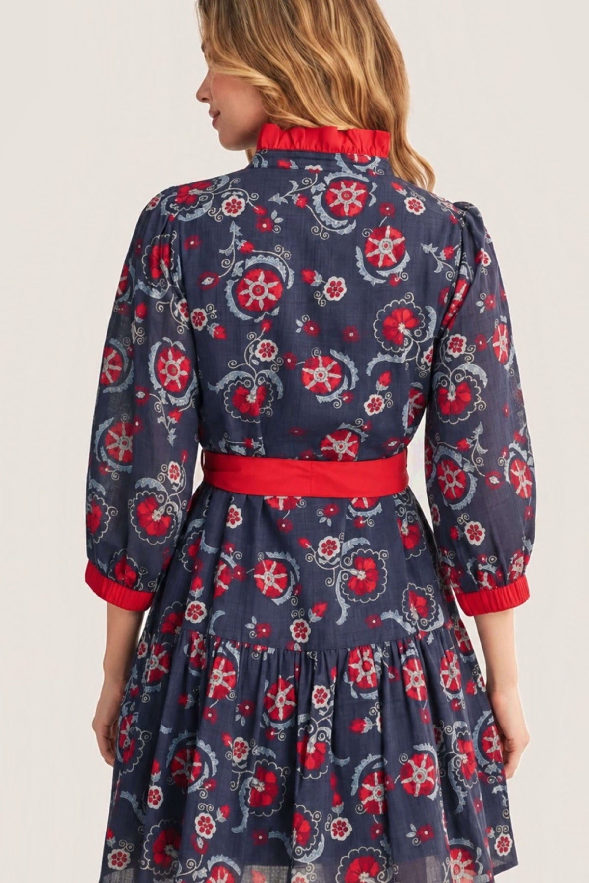 A woman shown from the back wearing a navy blue tiered dress with a red and white floral pattern, a red waist belt, and red trim on the collar and cuffs.