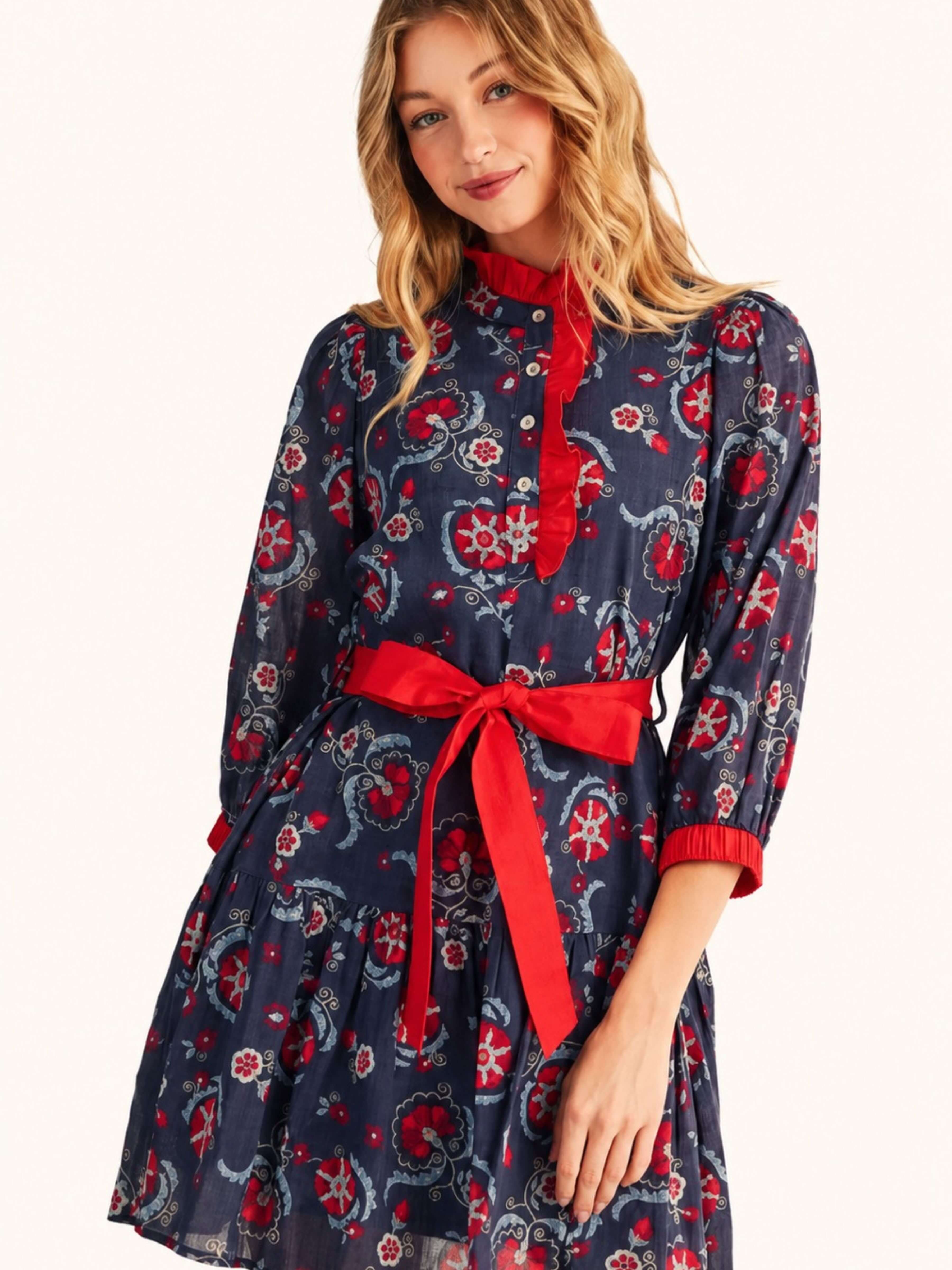 A woman modeling a navy blue long-sleeve dress with a red and white paisley pattern, featuring a red mock neck collar, red button placket, and a matching red waist tie belt.