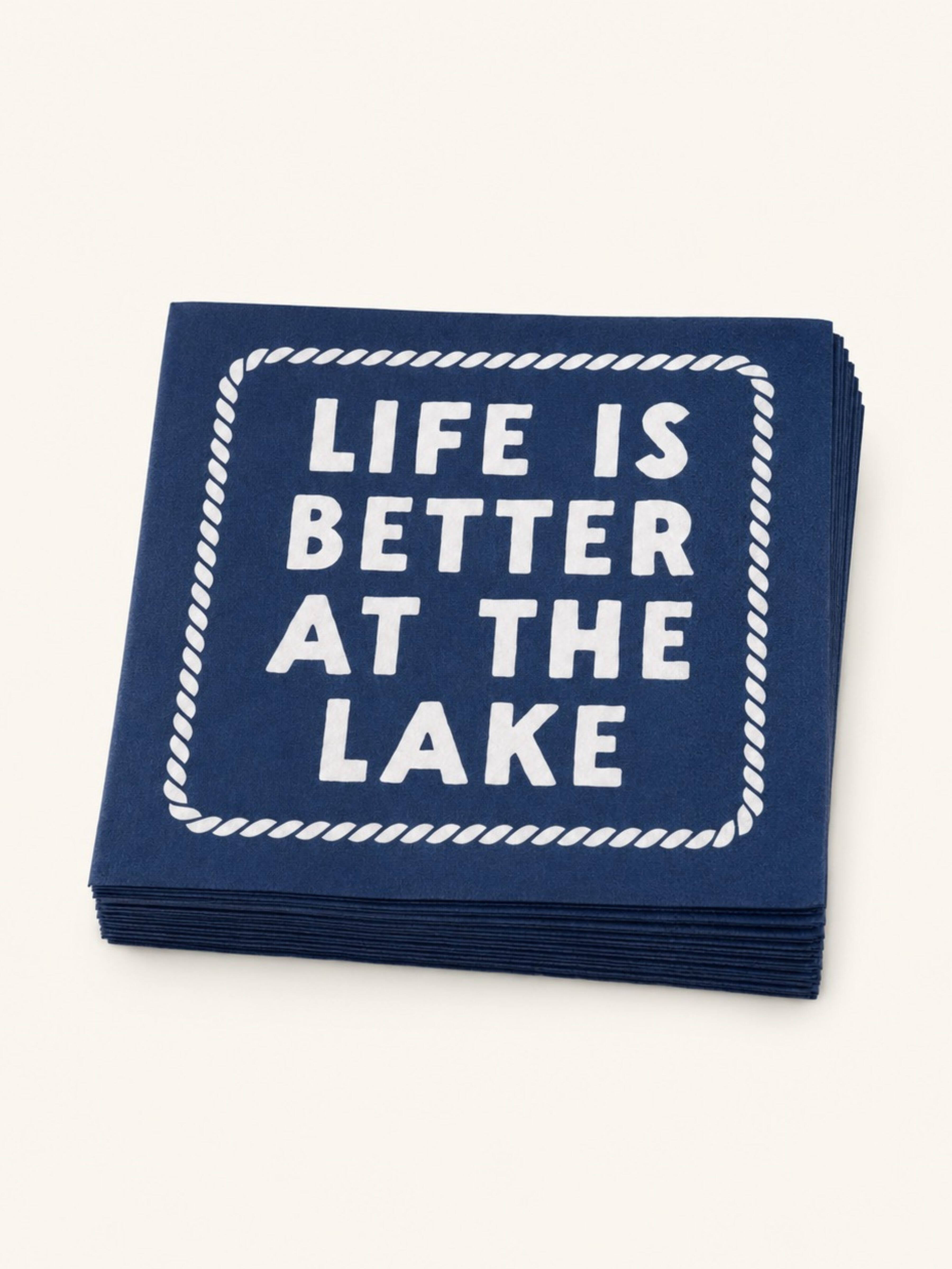 A stack of navy blue paper napkins with white printed text reading 'LIFE IS BETTER AT THE LAKE' inside a rope-style border, shown on a light cream background.
