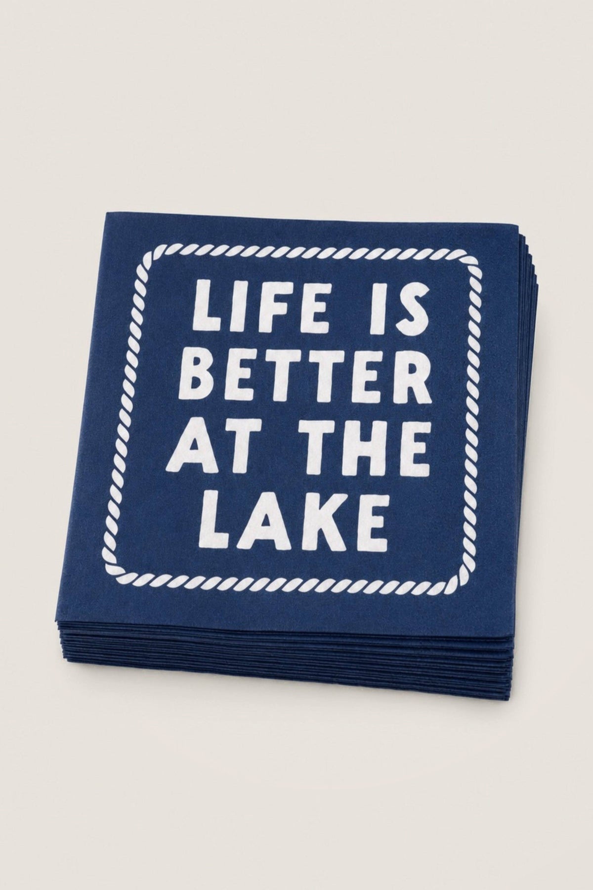 A stack of navy blue paper napkins with white printed text reading 'LIFE IS BETTER AT THE LAKE' inside a rope-style border, shown on a light cream background.