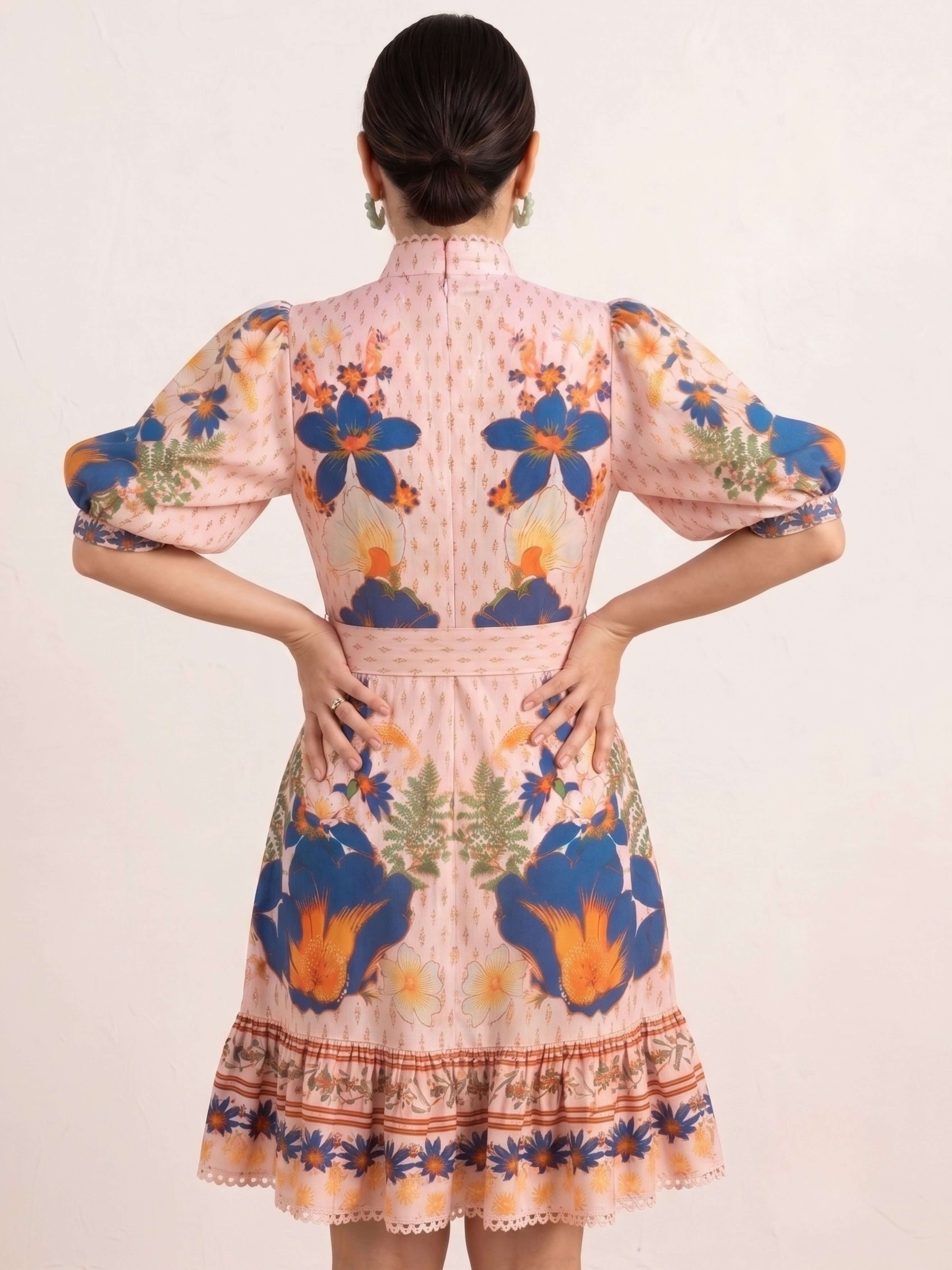 A woman viewed from the back wearing a short pink dress with puff sleeves and a colorful floral print in blue, orange, and green, set against a plain light beige background.