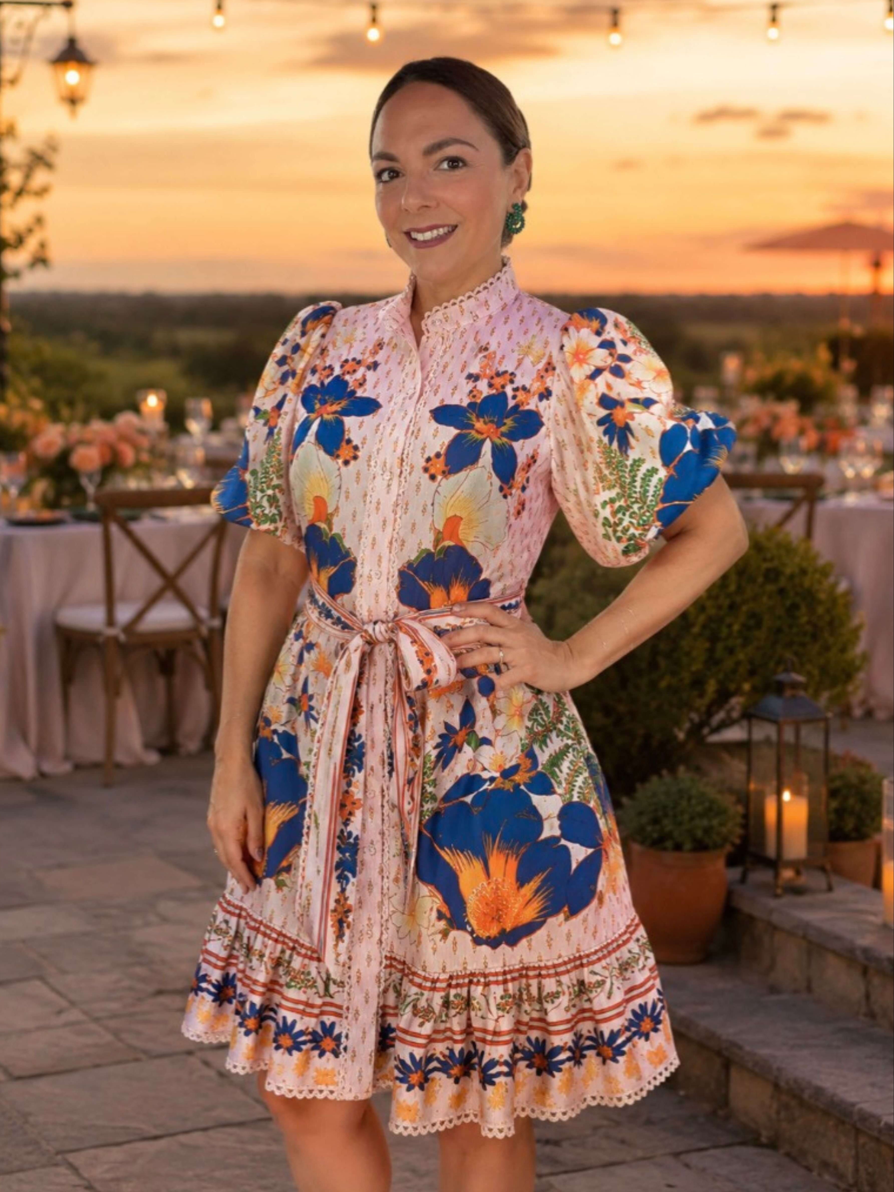 A woman wearing a colorful floral print dress with puff sleeves and a tie waist, standing outdoors at sunset on a patio with tables and string lights in the background.