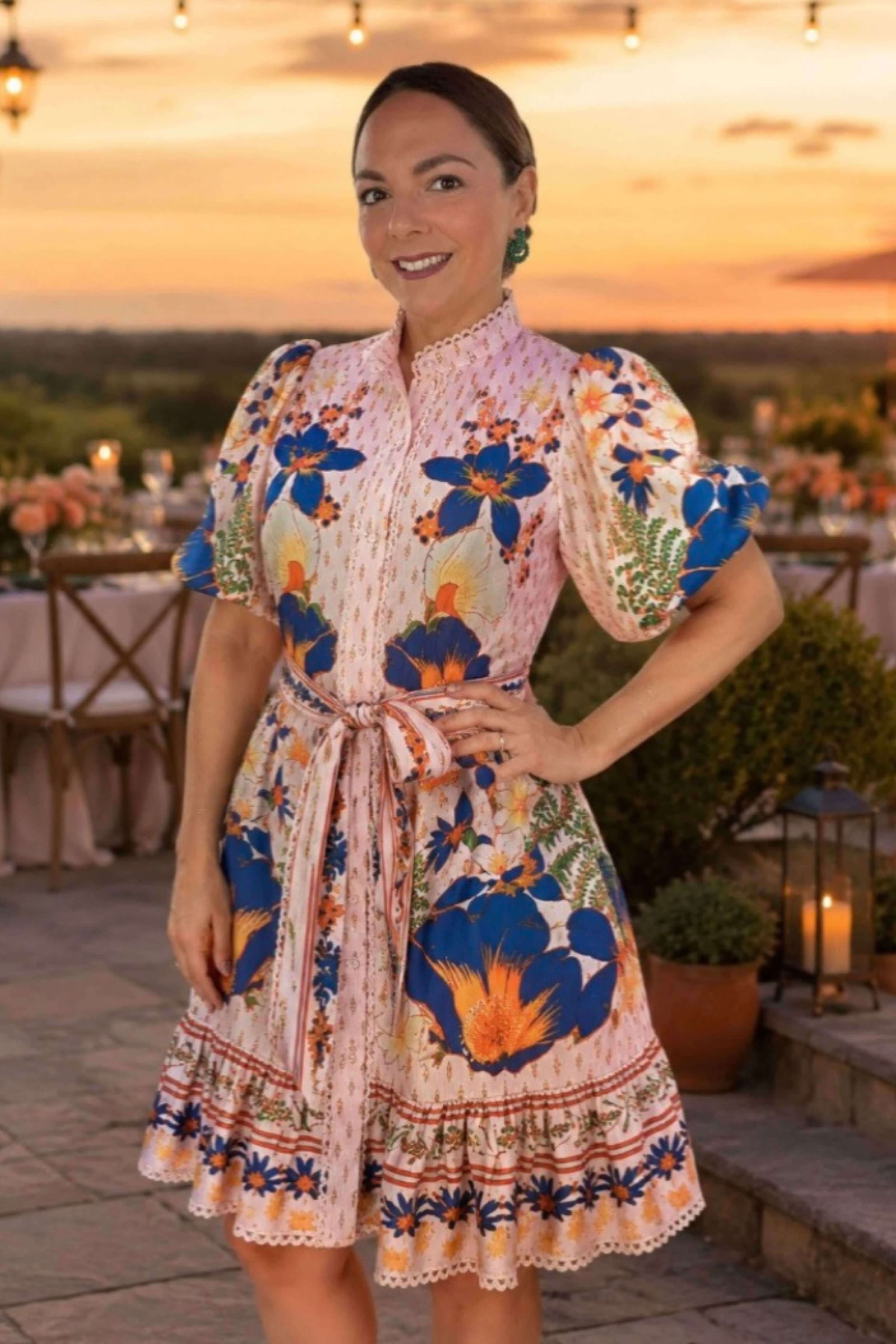 A woman wearing a short puff-sleeve midi dress with a colorful floral print, standing outdoors at sunset with tables and garden decor blurred in the background.