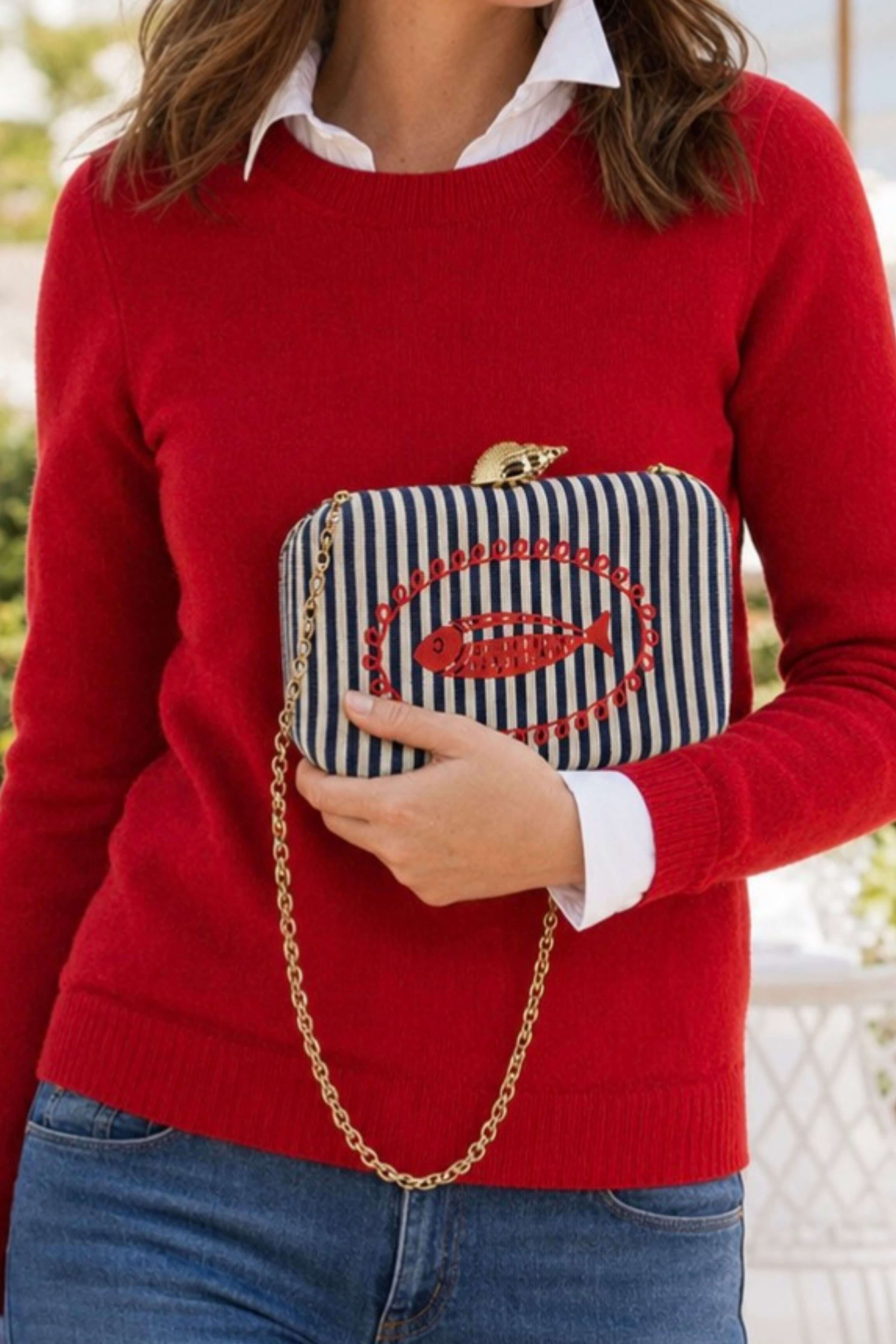 A woman wearing a red knit sweater and blue jeans holds a navy and white striped clutch bag with a red fish motif and a gold chain strap.