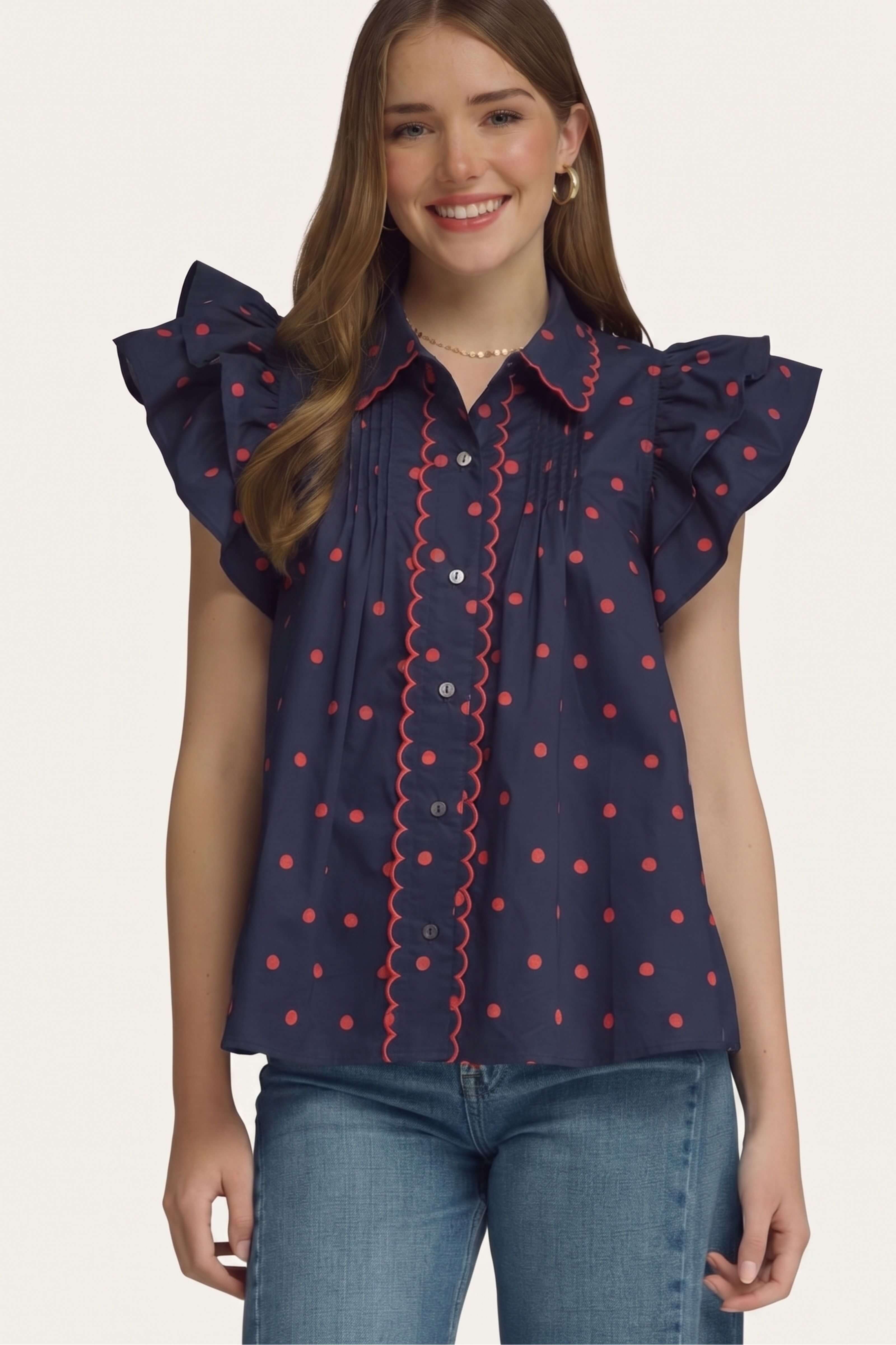A woman wearing a navy blue short-sleeve blouse with red polka dots, red scalloped trim, and ruffled shoulders, paired with blue jeans, against a plain light background.