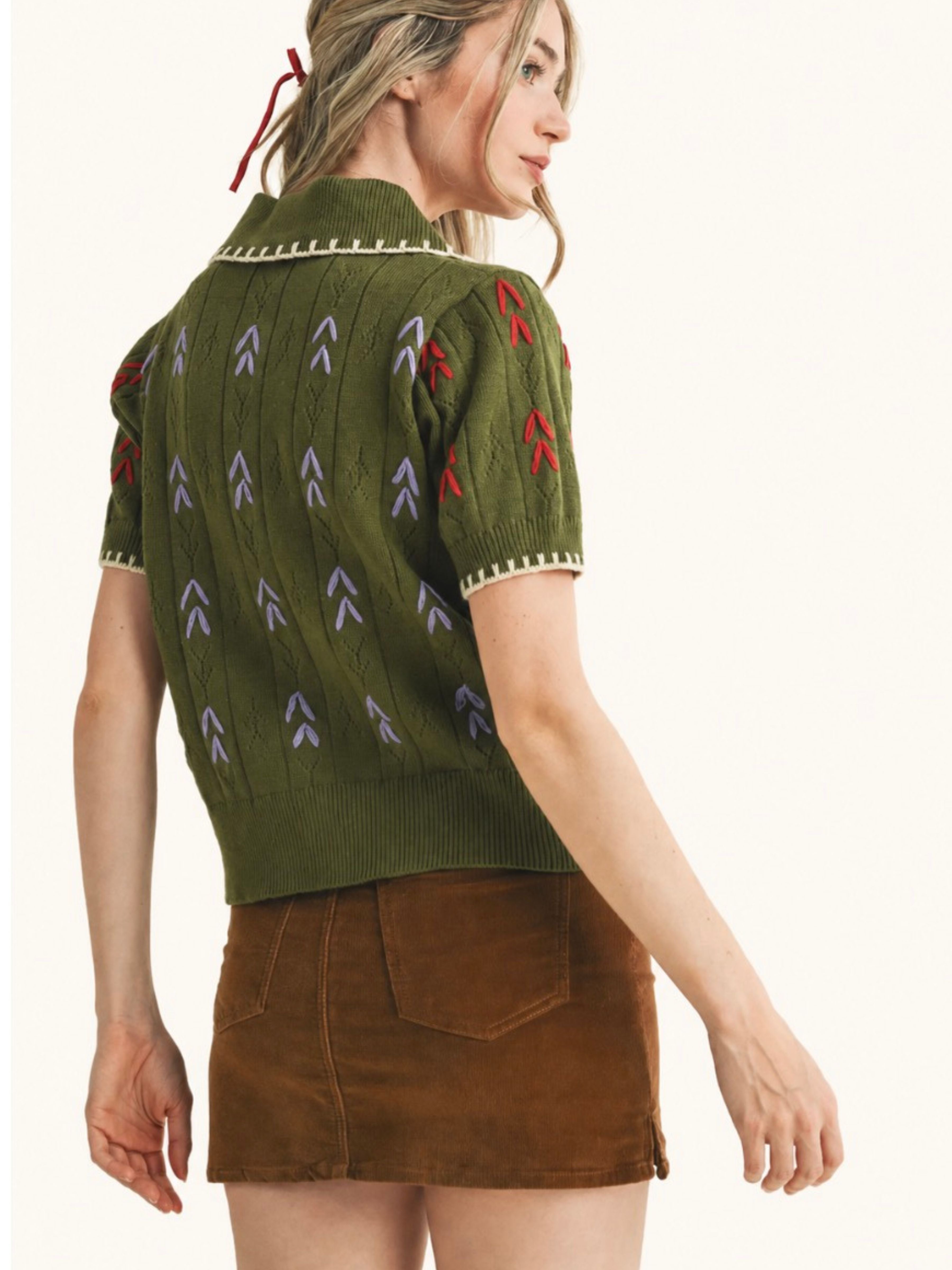 A woman shown from the back wearing a short-sleeve olive green knit sweater with red and gray chevron patterns and a brown mini skirt, against a plain light background.