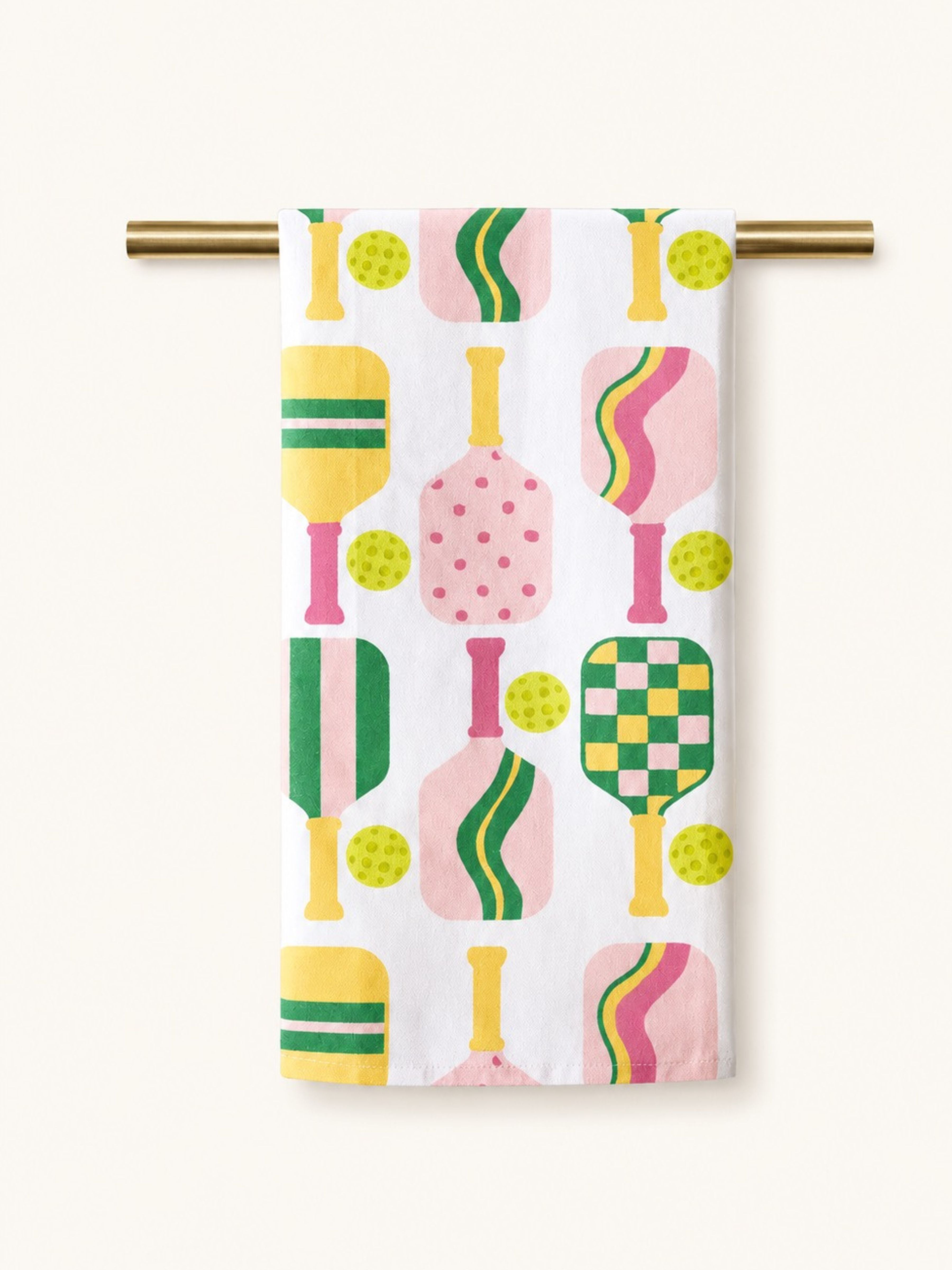 A white kitchen towel with a colorful retro pattern of tennis rackets, balls, and strokes, hanging from a gold metal rod against a light beige background.