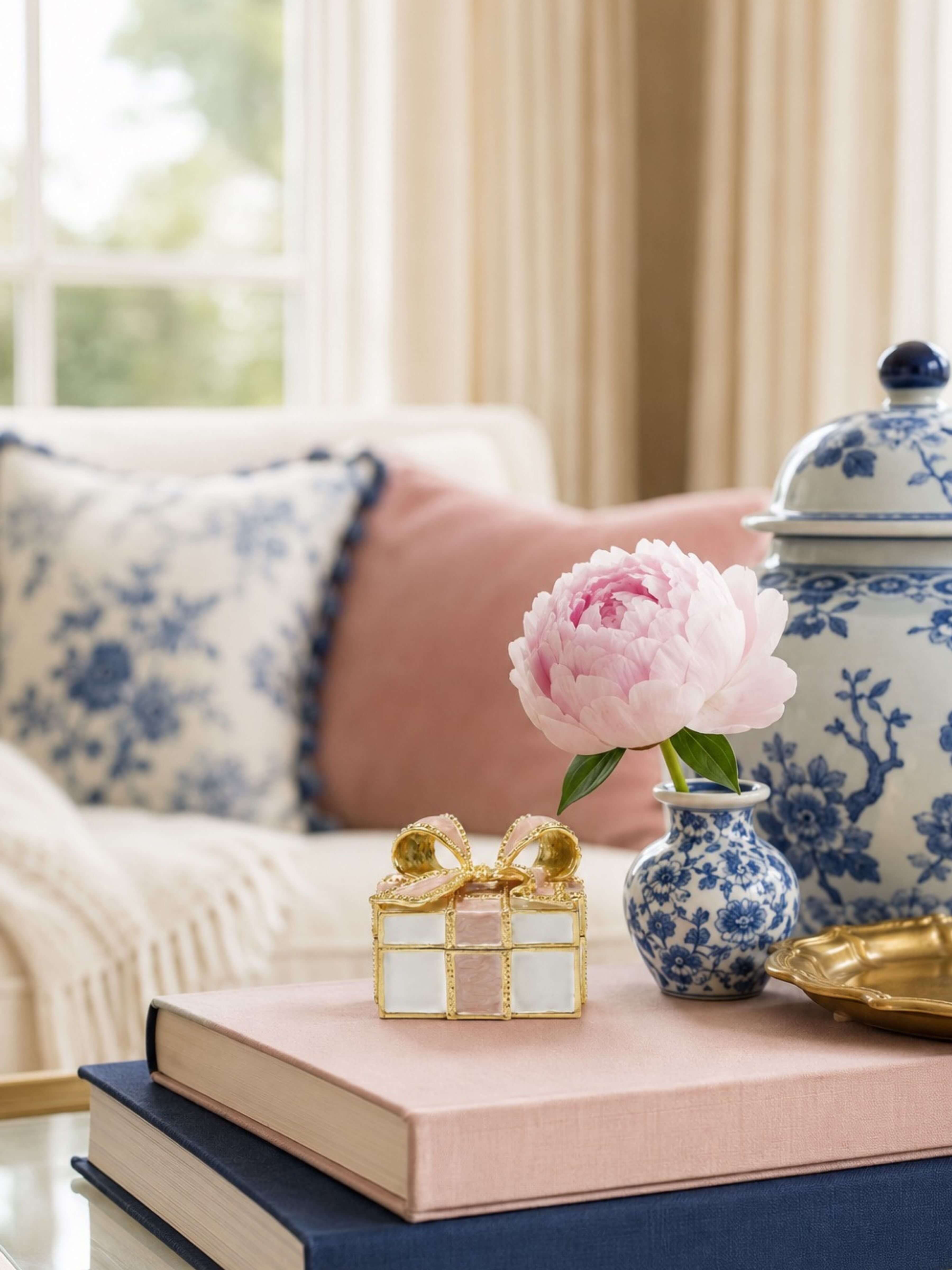 A styled interior scene with a pink peony in a blue-and-white ceramic vase, a small gold gift box, a pink book, and a navy book on a table in a softly lit room with patterned pillows and a blue-and-white lidded jar in the background.