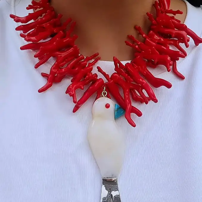 A close-up of a person wearing a chunky coral-style necklace in bright red, with a white bird pendant featuring a small red eye and a blue accent, over a white top.