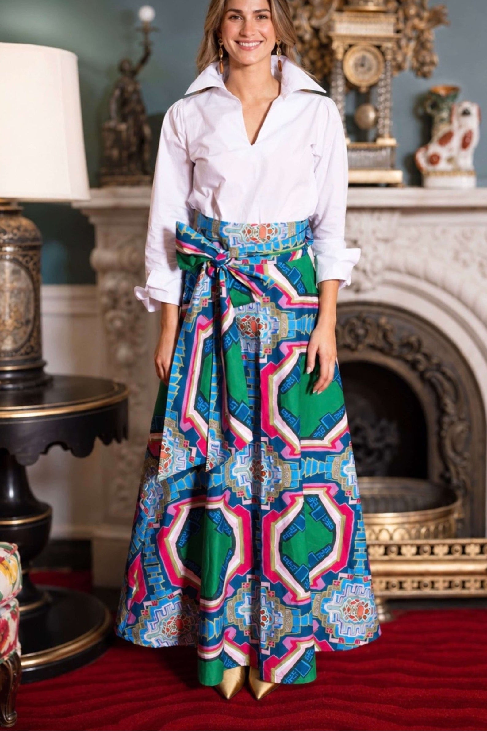 A woman stands in an ornate room wearing a white collared blouse and a long, wide skirt with a bold geometric pattern in green, blue, pink, and white. The skirt has a matching fabric tie belt at the waist.