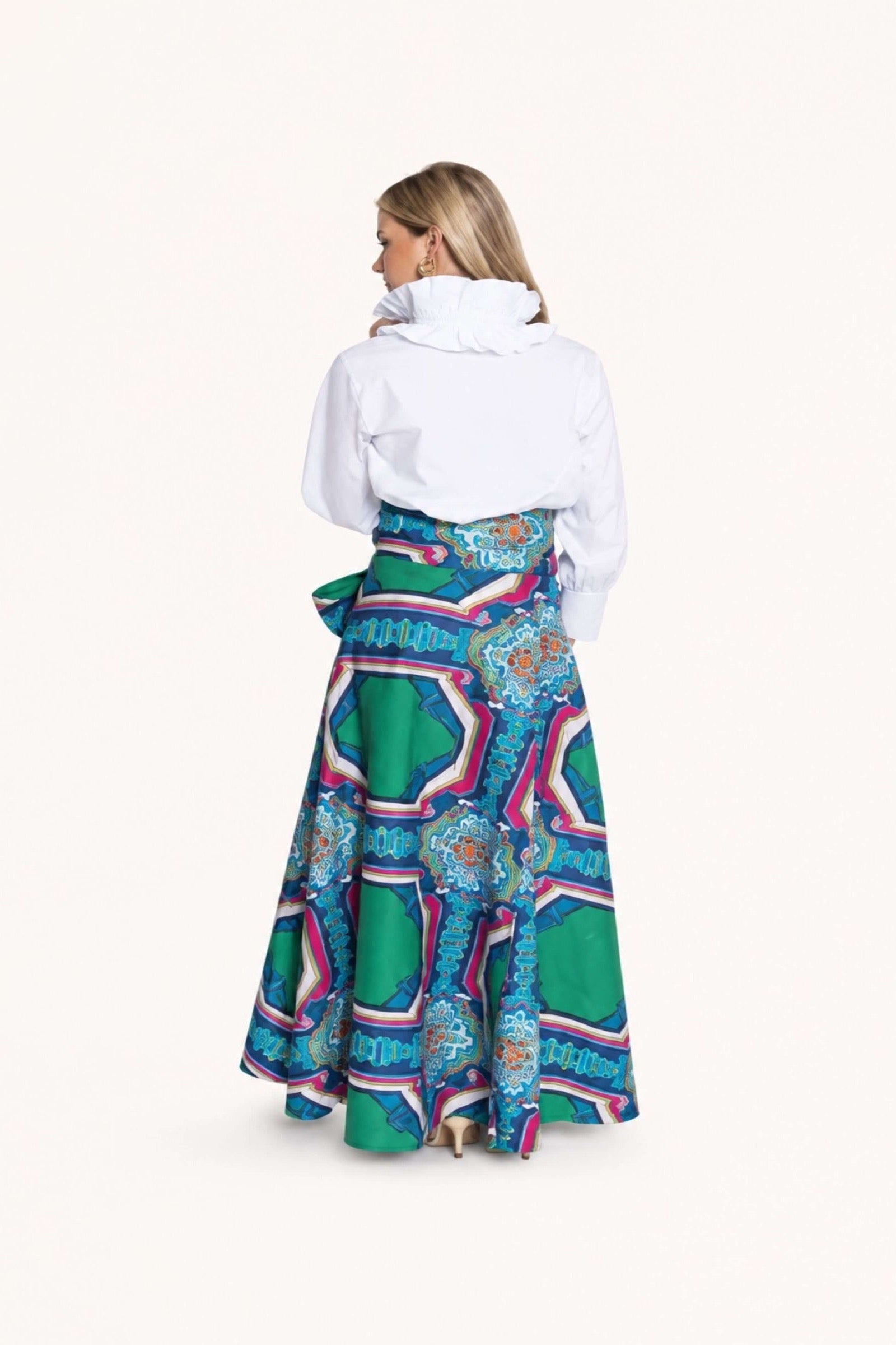 A woman shown from the back wearing a white long-sleeve top and a full, flared skirt with a bold geometric and floral pattern in green, blue, pink, and purple, against a plain off-white studio background.