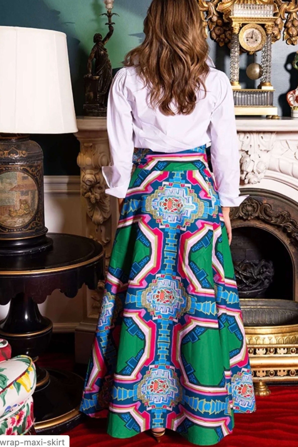 A woman shown from behind wearing a white long-sleeve blouse and a high-waisted, full-length A-line skirt with a vibrant, multicolored geometric and medallion pattern in green, blue, pink, and orange. She is standing in an ornate, vintage-style room with a fireplace and antique furniture.