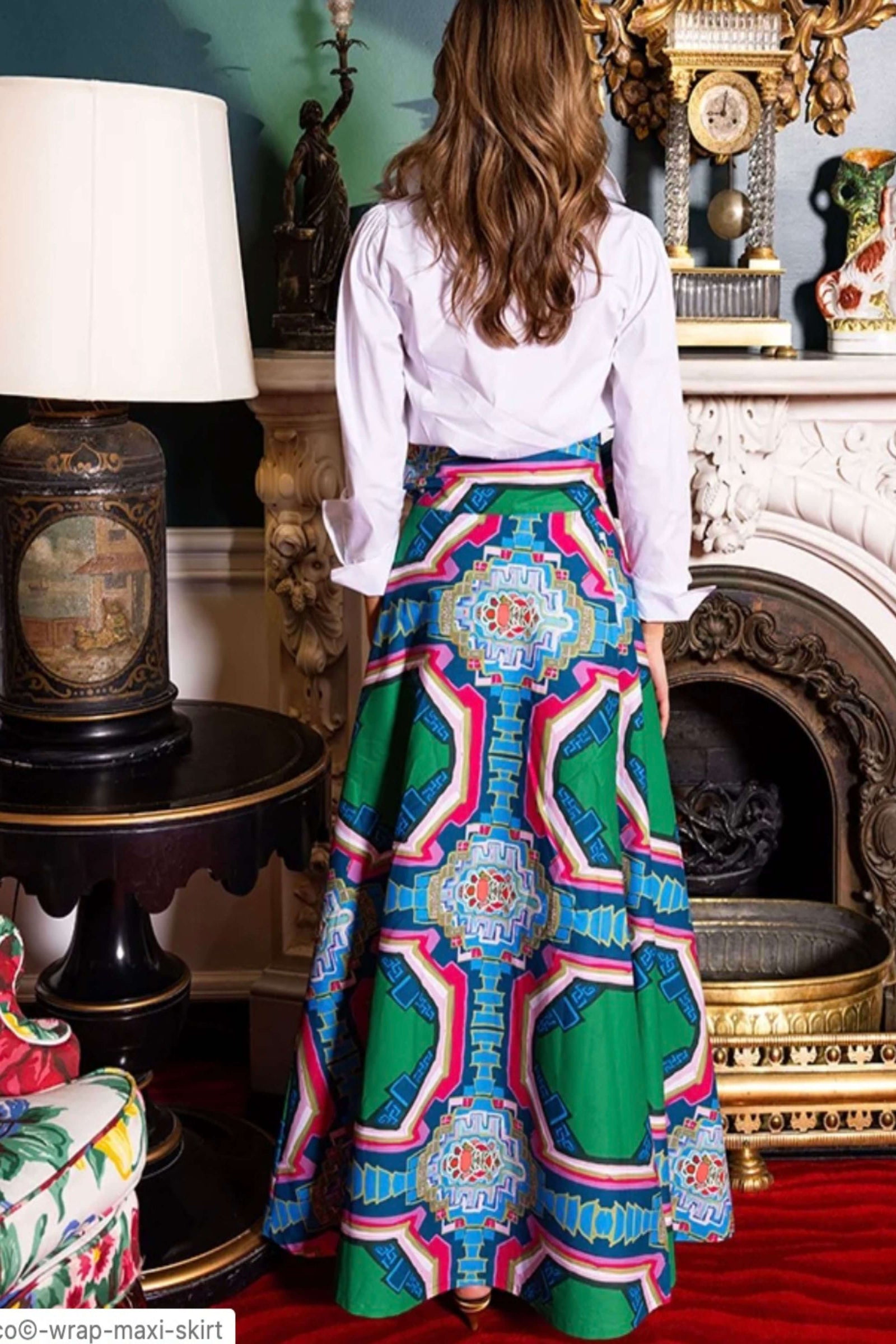 A woman shown from behind wearing a white blouse and a full, flared maxi skirt with a bold geometric pattern in green, blue, pink, and orange. She is standing in an ornate room with a fireplace, lamp, and decorative furniture.