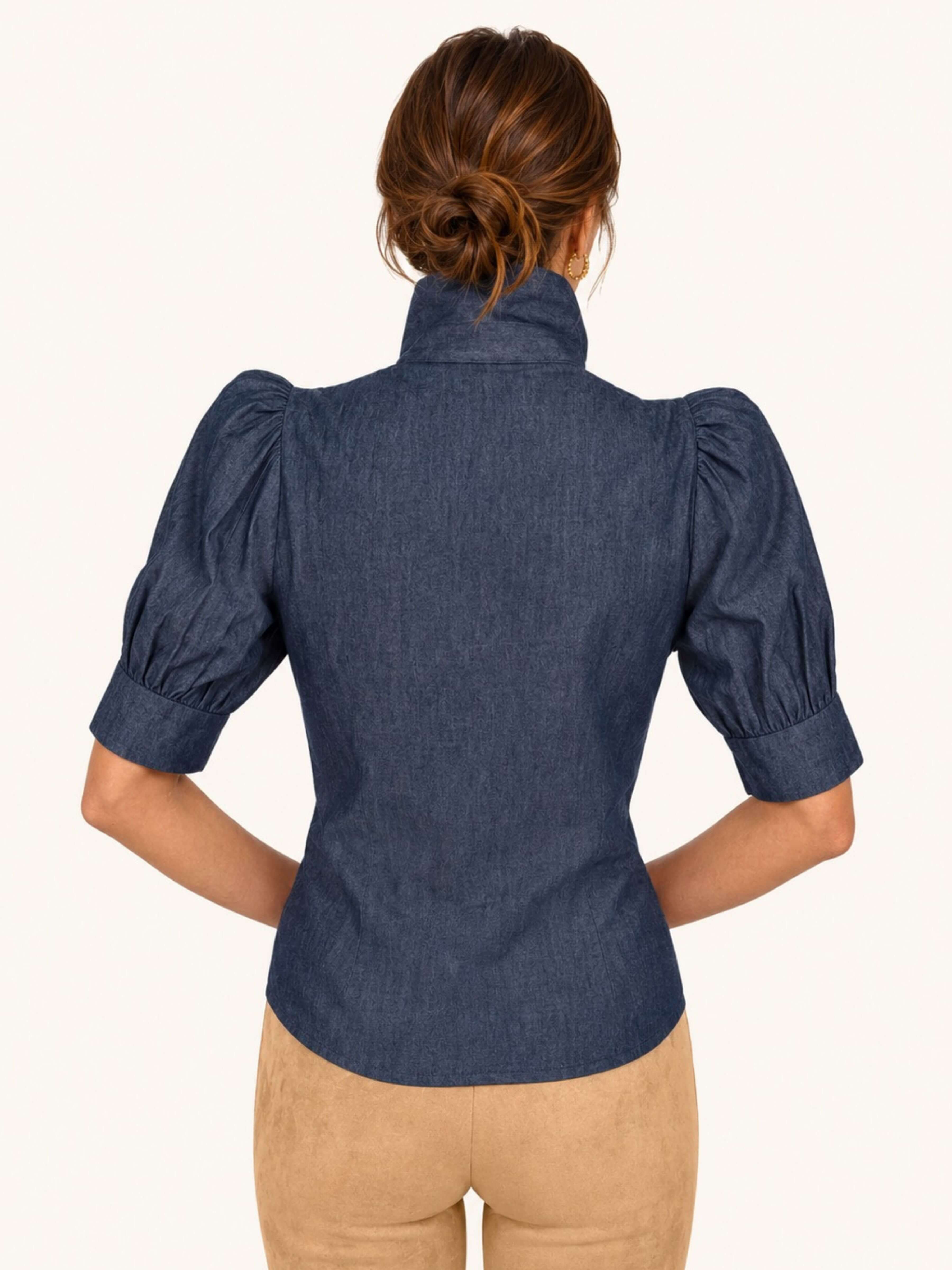 A woman shown from the back wearing a navy blue short-sleeve blouse with puffed sleeves and a high mock neck, paired with tan pants, against a plain off-white background.