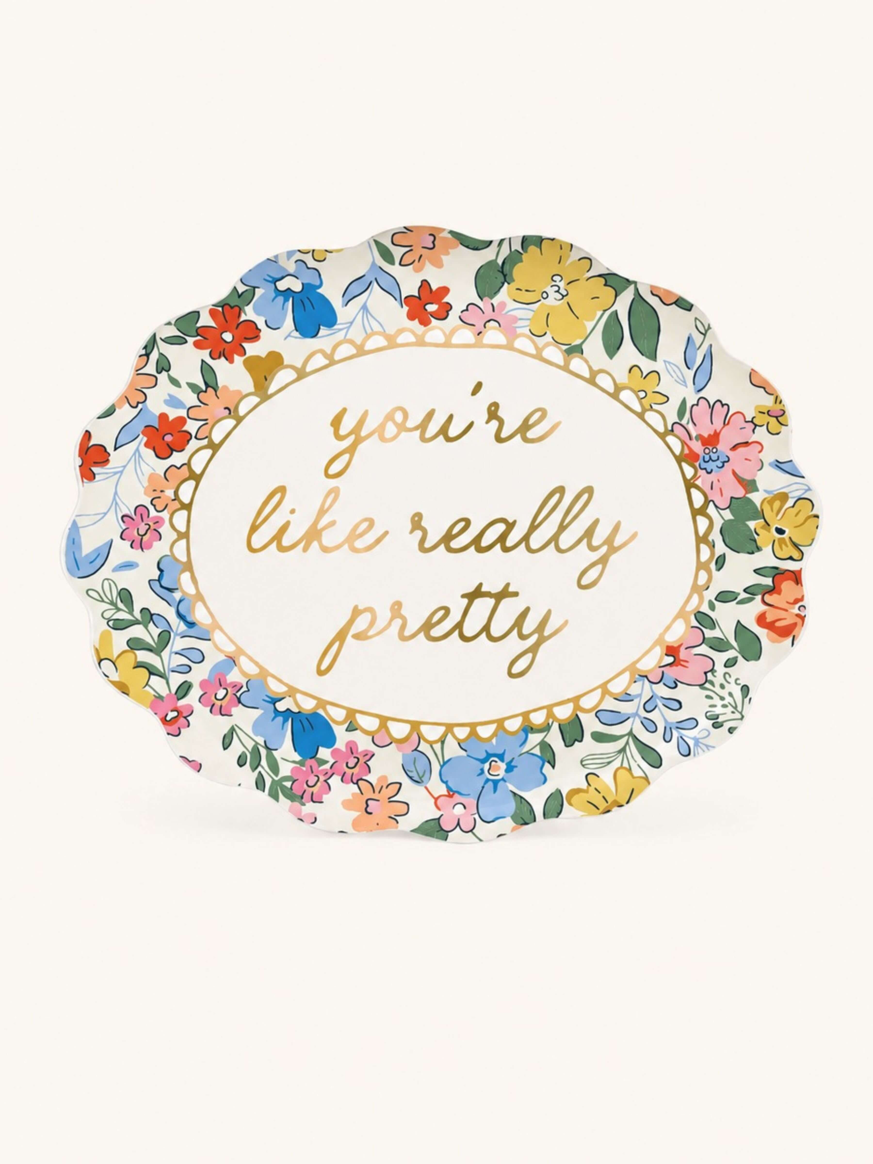 An oval paper plate with a scalloped edge, featuring a colorful floral border and the phrase 'you're like really pretty' in gold cursive text in the center, set against a plain off-white background.