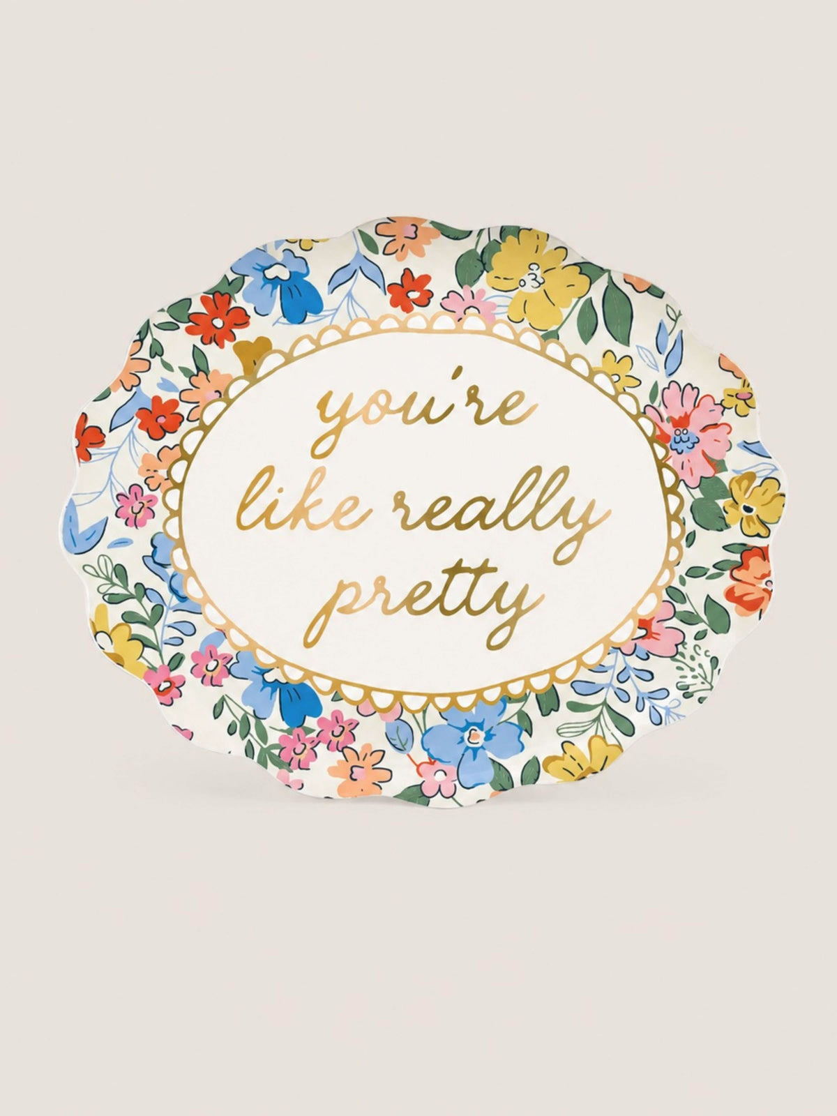 An oval paper plate with a scalloped edge, featuring a colorful floral border and the phrase 'you're like really pretty' in gold cursive text in the center, set against a plain off-white background.