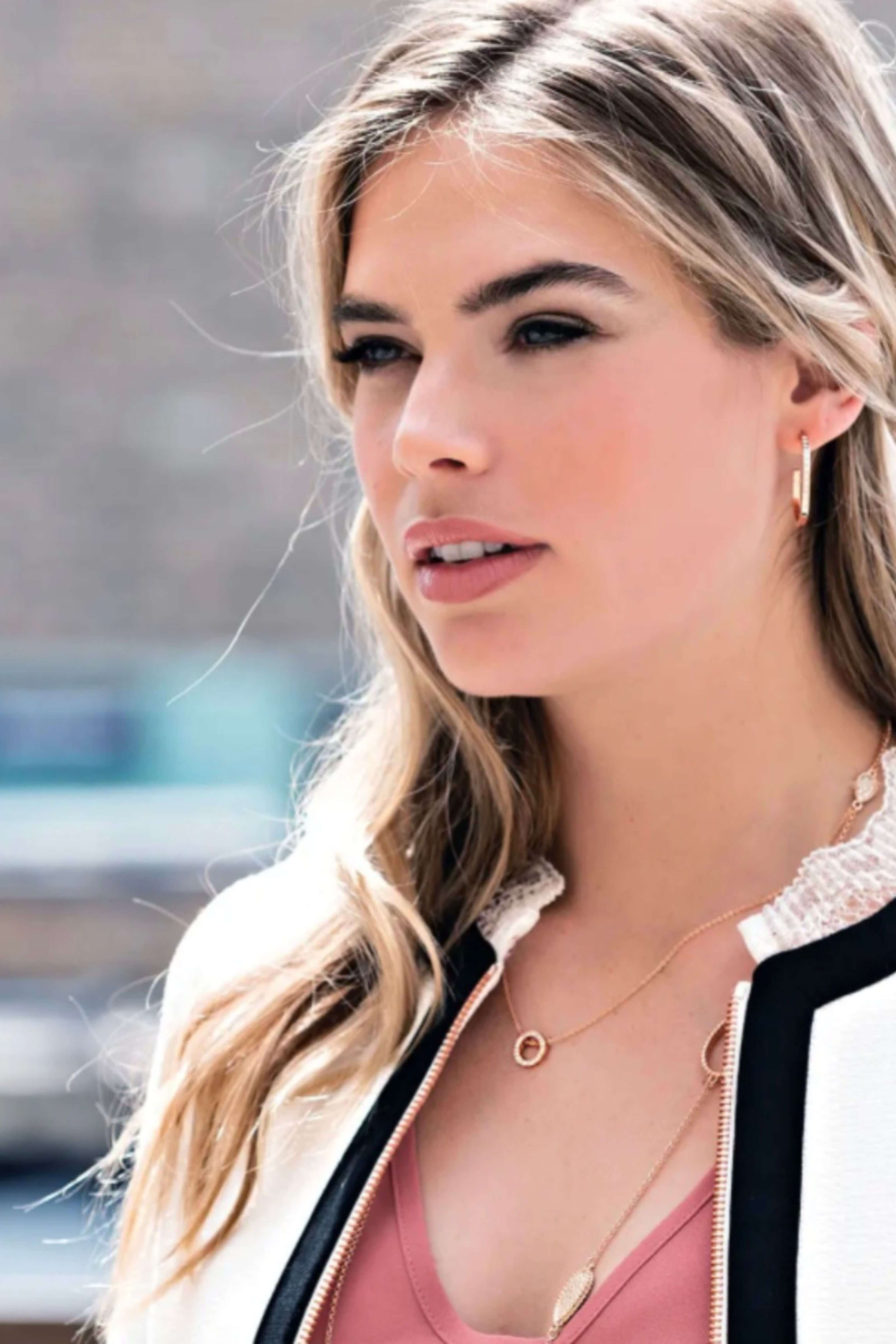 A close-up lifestyle shot of a woman with blonde hair wearing a delicate gold necklace with a circular pendant and a small hoop earring, paired with a pink top and a white jacket with black trim.