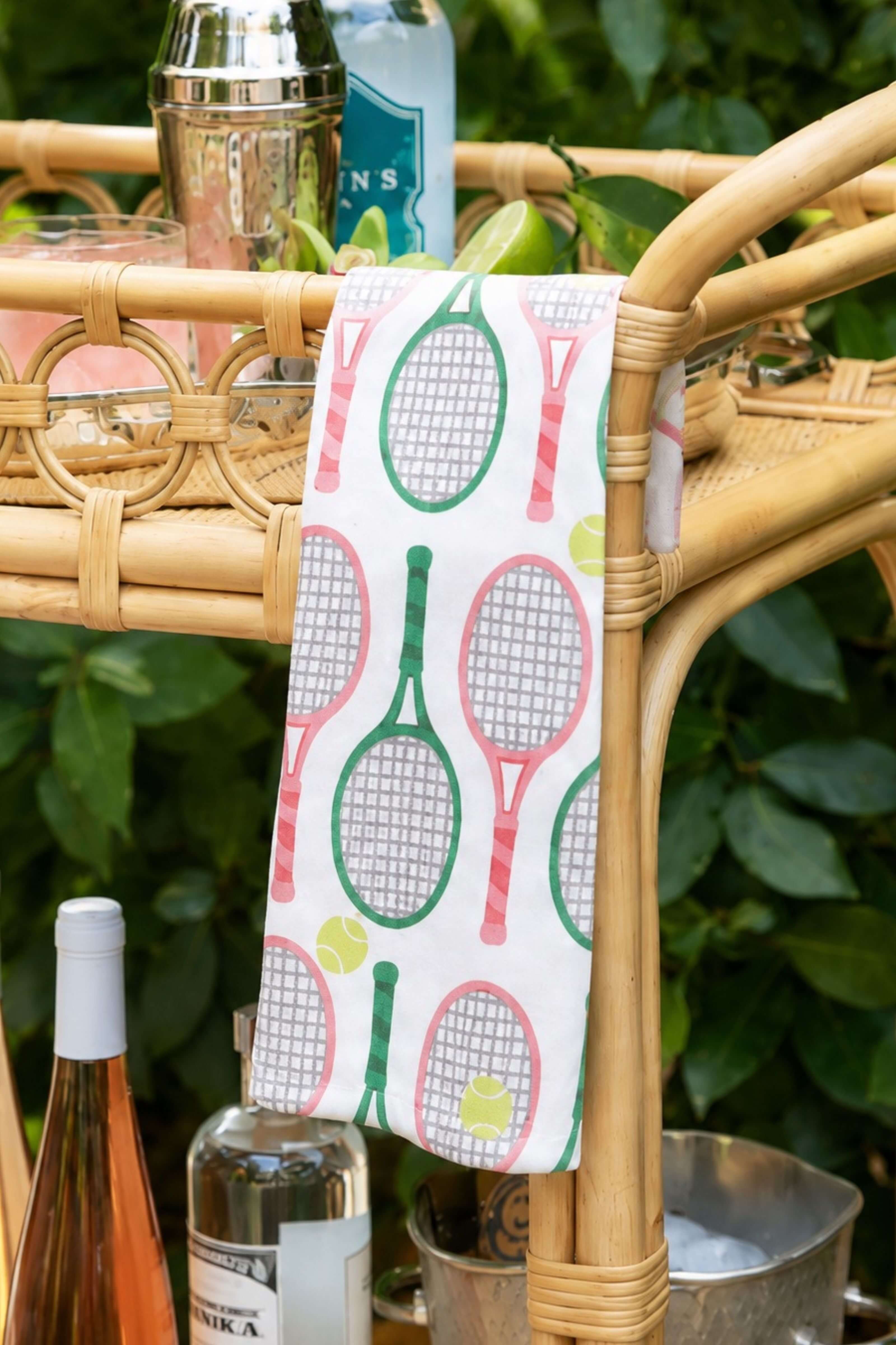 A white kitchen towel with a colorful tennis racket pattern hangs from the arm of a bamboo bar cart, styled with drinks and limes against a green leafy background.