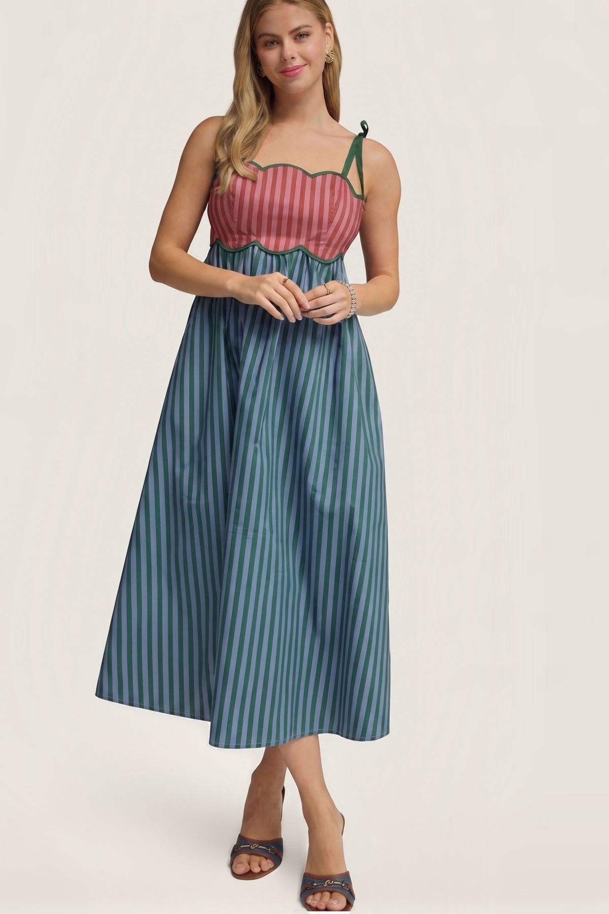 A woman modeling a sleeveless midi dress with a pink and red striped bodice and teal and blue vertical striped skirt, paired with brown heeled sandals, against a light beige studio background.