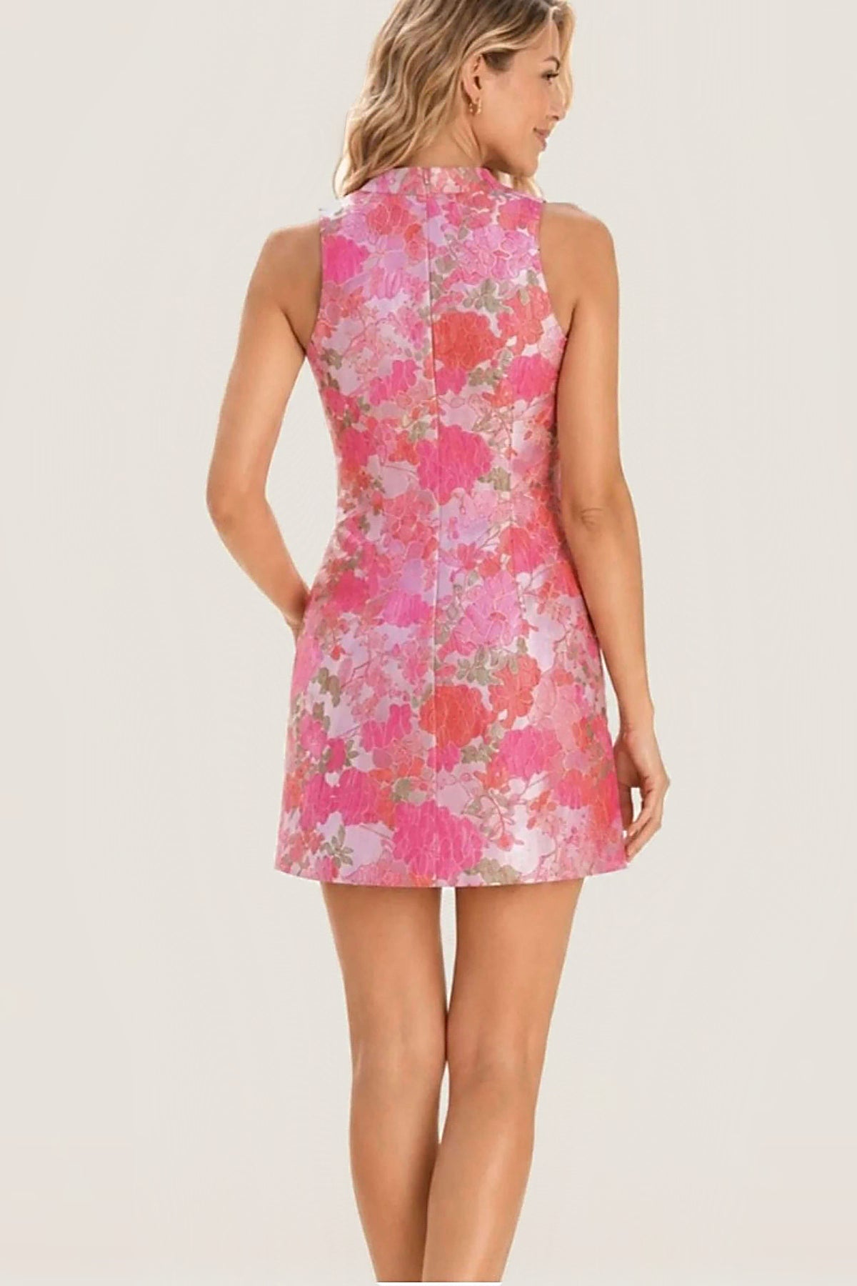 A woman shown from the back wearing a sleeveless high-neck mini dress with a pink, coral, and white floral pattern on a light background.