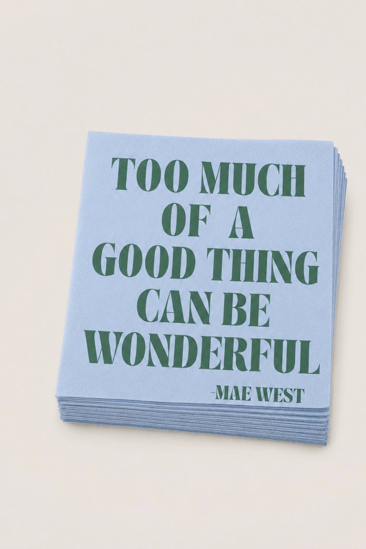 A stack of light blue paper napkins featuring the quote 'TOO MUCH OF A GOOD THING CAN BE WONDERFUL -MAE WEST' printed in dark green serif lettering, set against a solid off-white background.