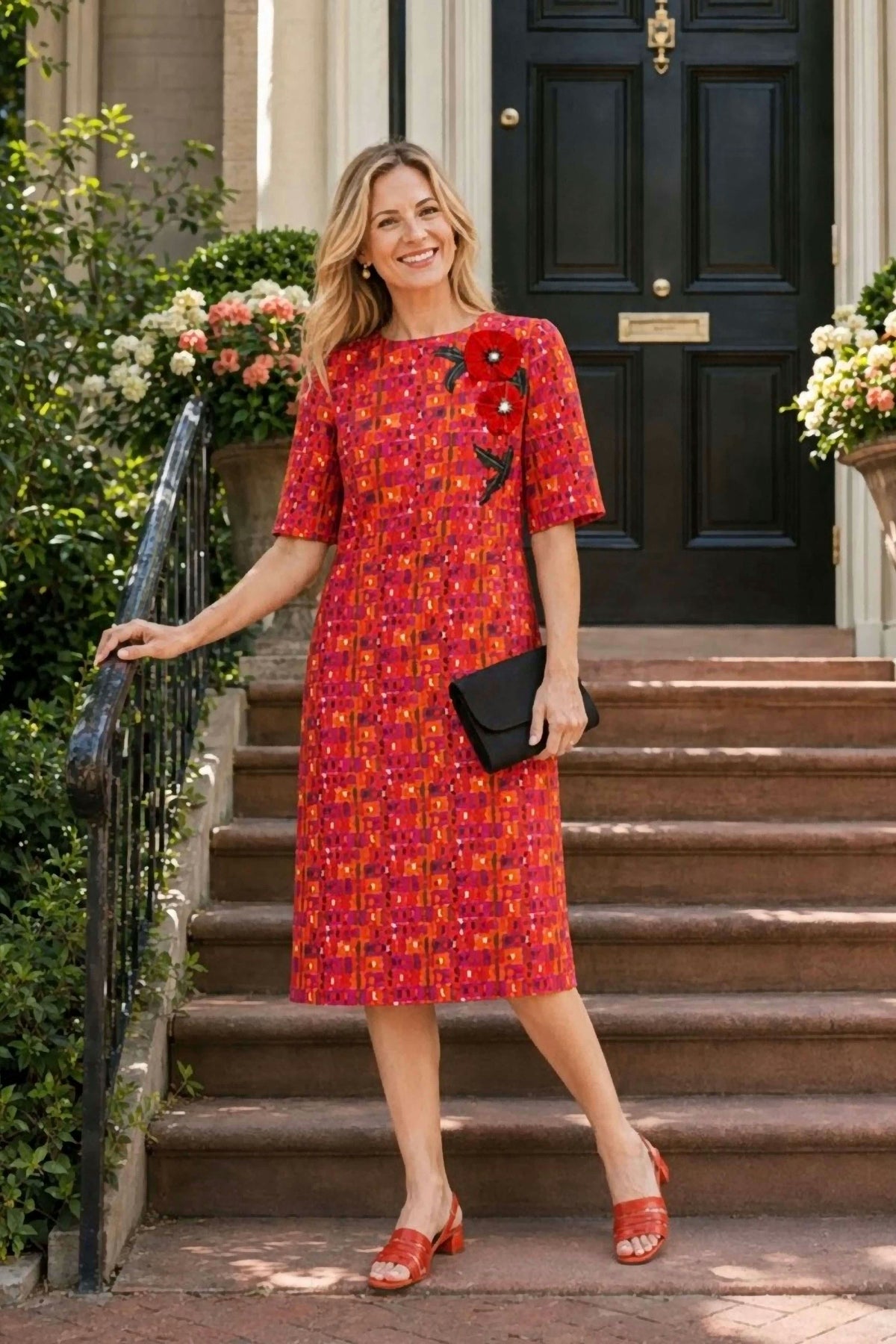A woman stands on stone steps in front of a building entrance wearing a short-sleeve knee-length dress with an allover red, orange, and yellow floral pattern, plus a black clutch and red sandals.