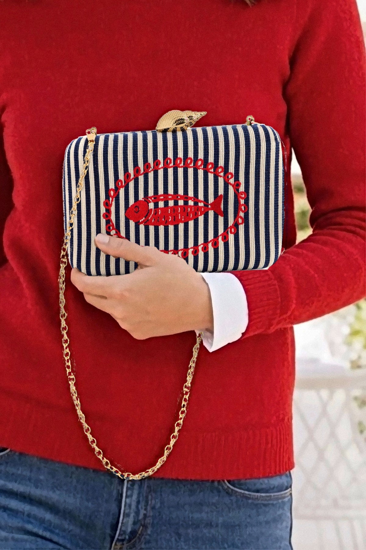 The Navy Marina Minaudière held in one hand with its gold chain hanging, worn with a red sweater and jeans.