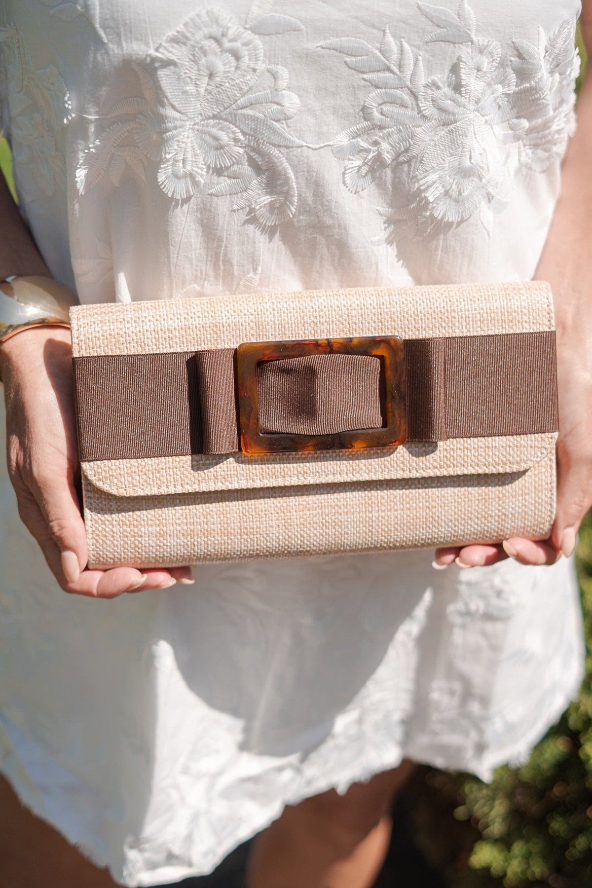 The Avery straw clutch held in both hands with a white embroidered dress.